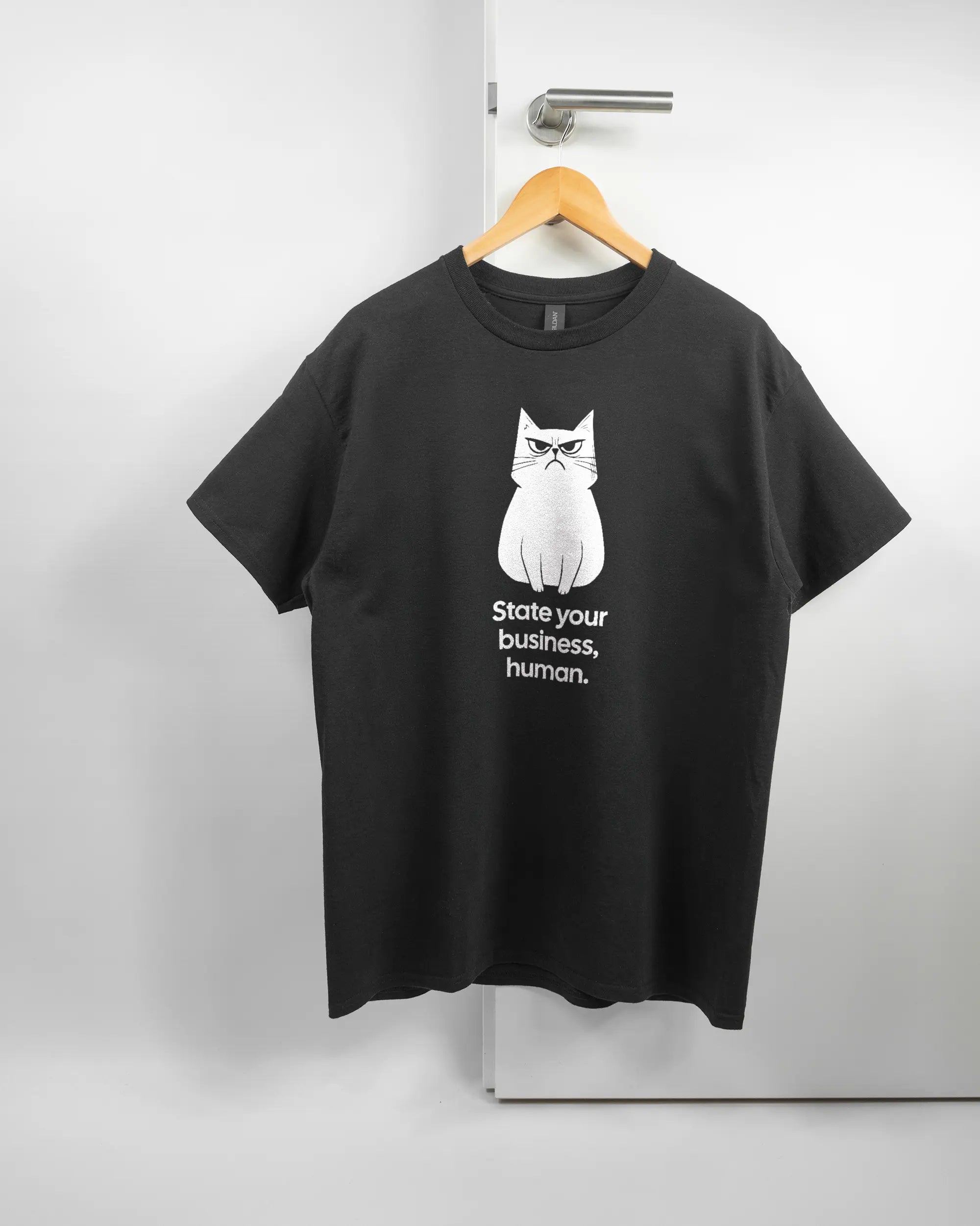 Vorderseite des Basic Unisex T-Shirt Regular von Maondo mit dem Design Grumpy Purr in der Farbe Black und in der Größe L auf einem Bügel an der Türklinke aufgehängt