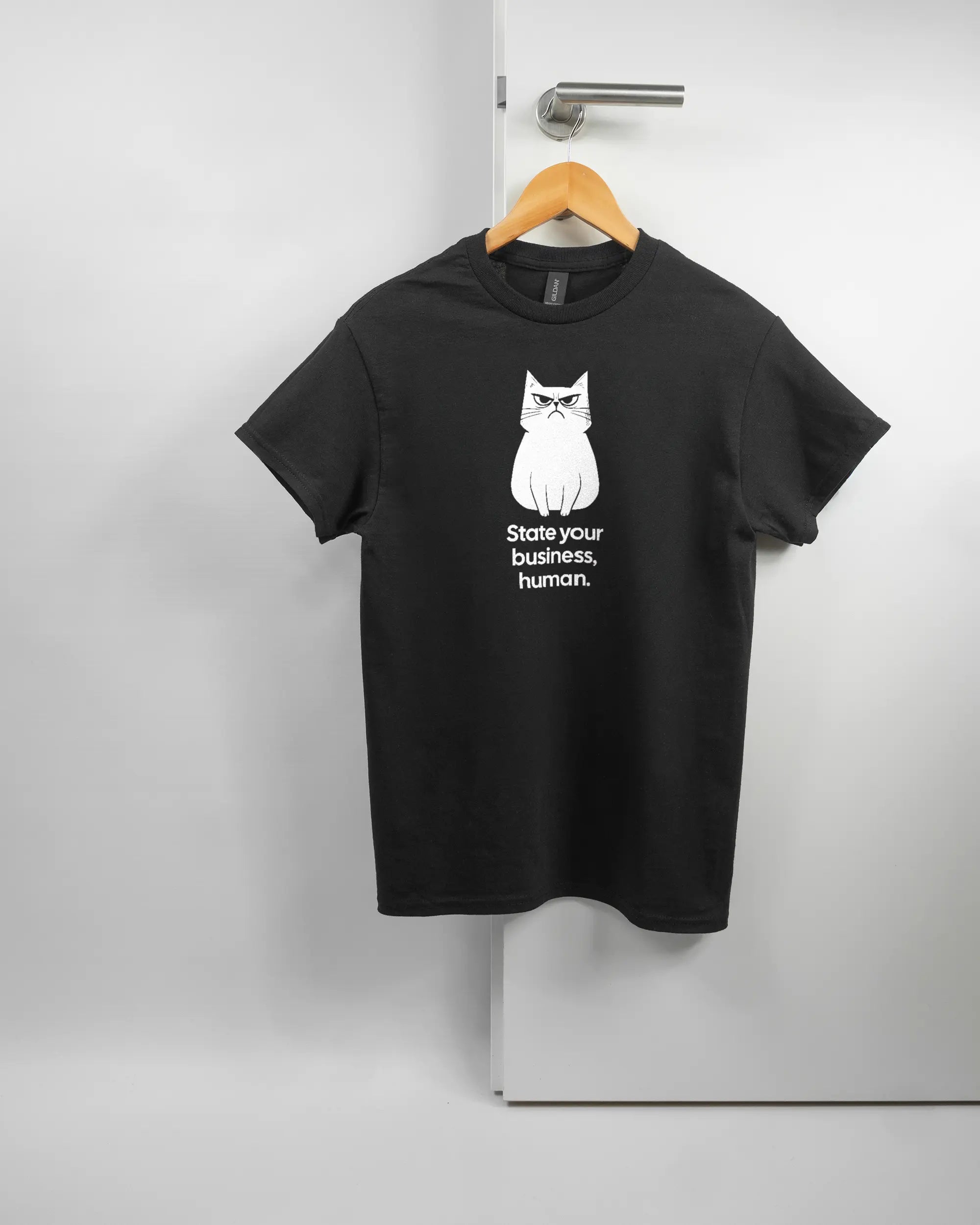 Vorderseite des Basic Unisex T-Shirt Regular von Maondo mit dem Design Grumpy Purr in der Farbe Black und in der Größe S auf einem Bügel an der Türklinke aufgehängt