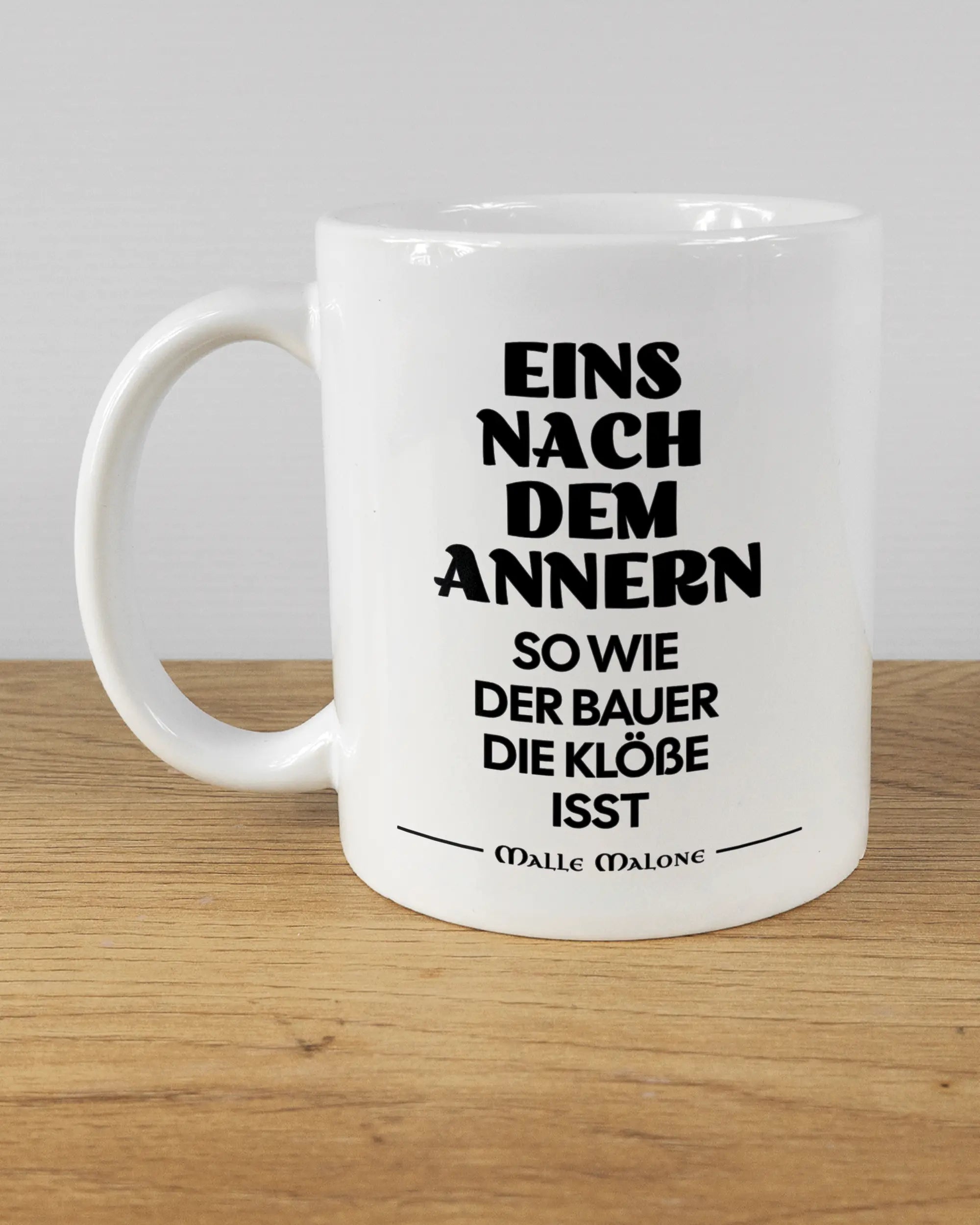 Titelbild, das die linke Seite der beidseitig bedruckten Keramiktasse von Maondo mit dem Design Eins nach dem Annern in der Farbe White auf einem Holztisch zeigt