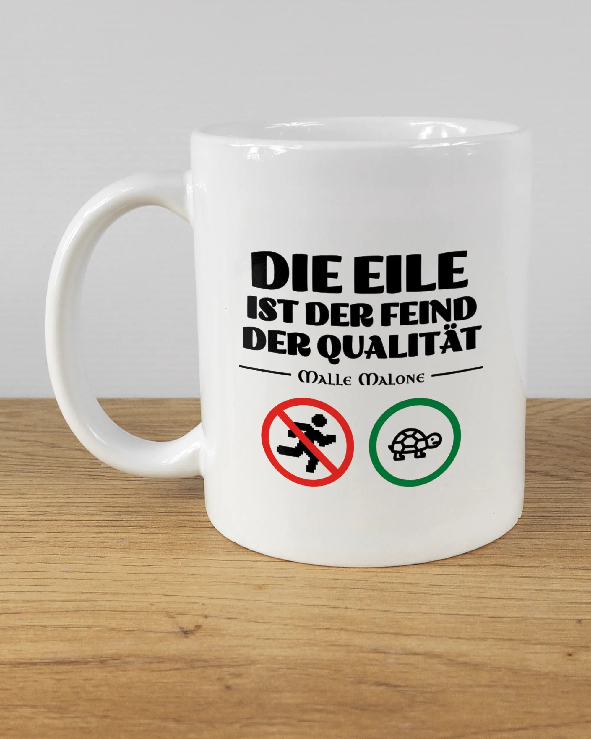 Titelbild, das die linke Seite der beidseitig bedruckten Keramiktasse von Maondo mit dem Design Keine Eile in der Farbe White auf einem Holztisch zeigt