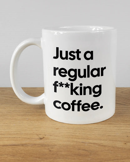 Titelbild, das die linke Seite der beidseitig bedruckten Keramiktasse von Maondo mit dem Design Regular Coffee in der Farbe White auf einem Holztisch zeigt