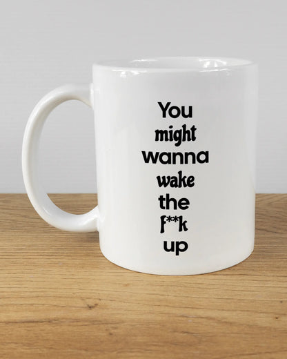 Titelbild, das die linke Seite der beidseitig bedruckten Keramiktasse von Maondo mit dem Design Wanna Wake in der Farbe White auf einem Holztisch zeigt