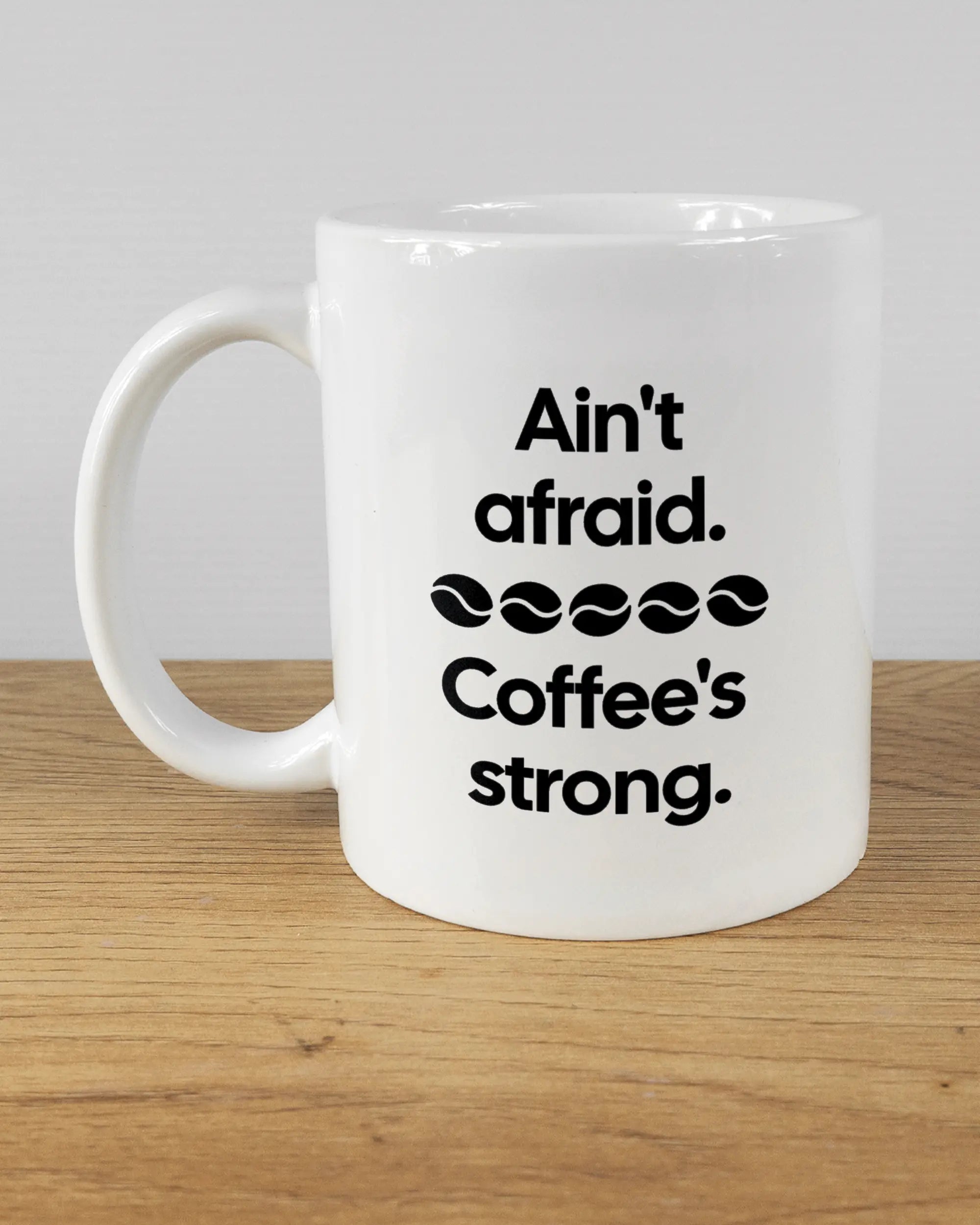 Titelbild, das die linke Seite der beidseitig bedruckten Keramiktasse von Maondo mit dem Design Coffee is strong in der Farbe White auf einem Holztisch zeigt