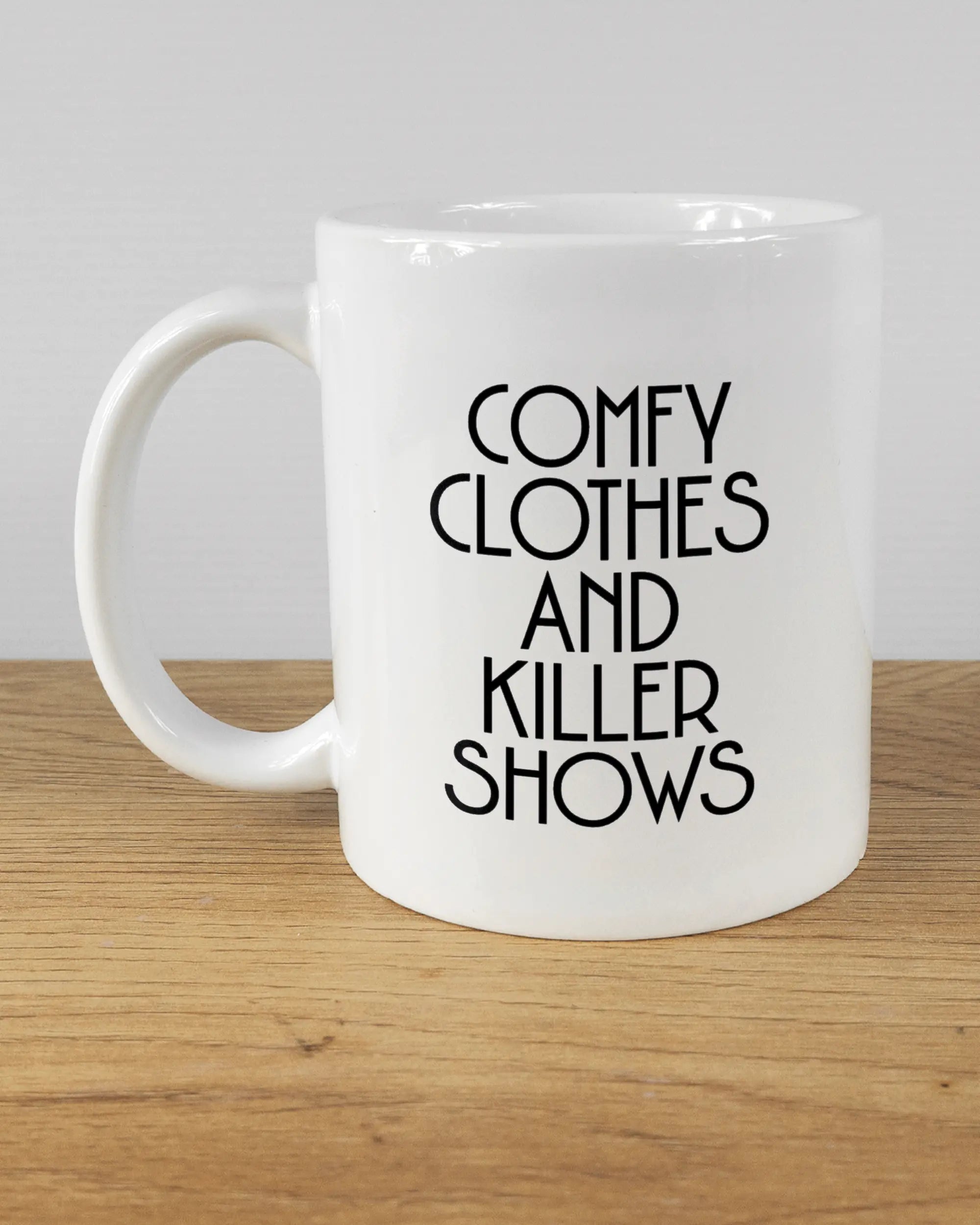 Titelbild, das die linke Seite der beidseitig bedruckten Keramiktasse von Maondo mit dem Design Killer Shows in der Farbe White auf einem Holztisch zeigt