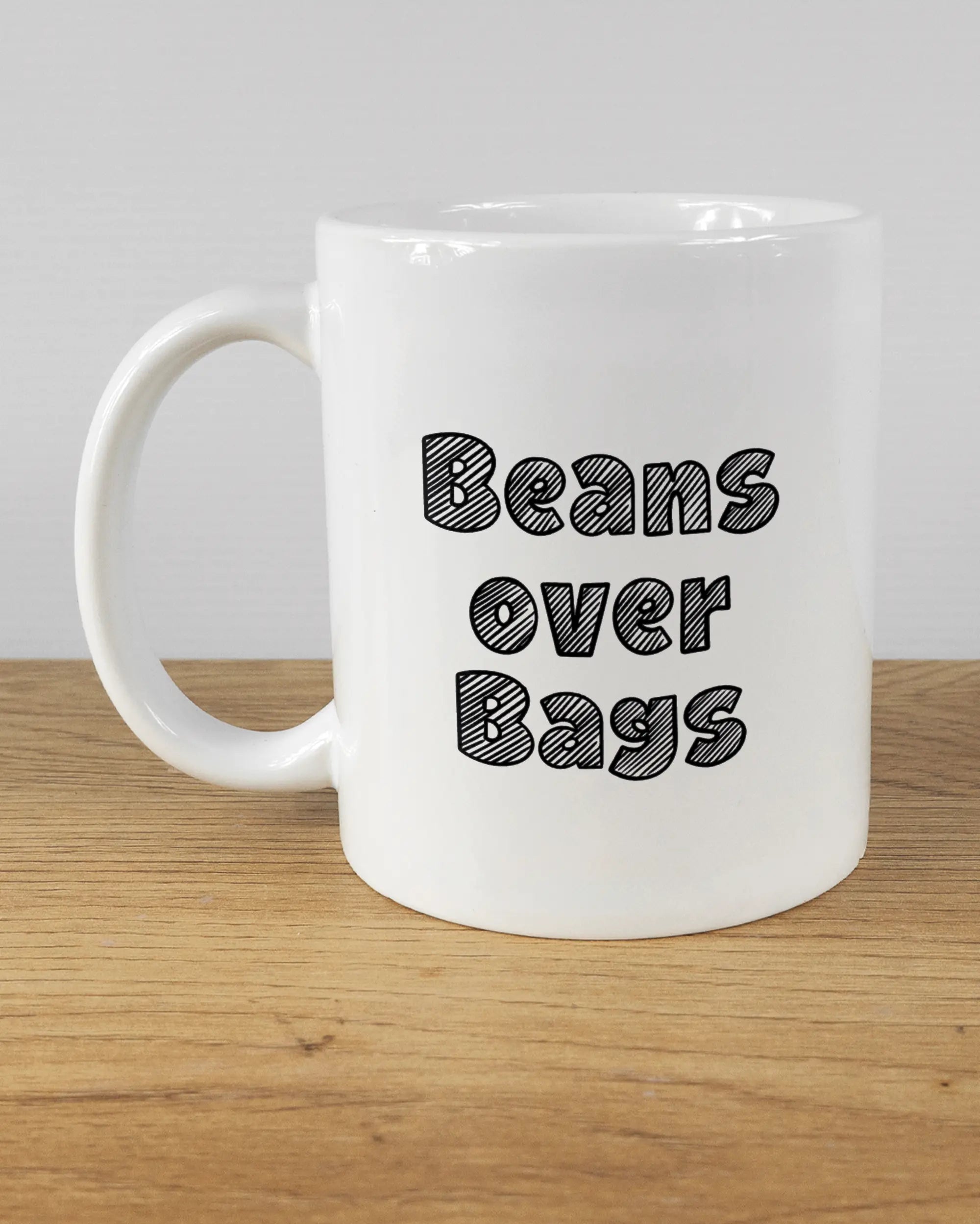 Titelbild, das die linke Seite der beidseitig bedruckten Keramiktasse von Maondo mit dem Design Beans over Bags in der Farbe White auf einem Holztisch zeigt