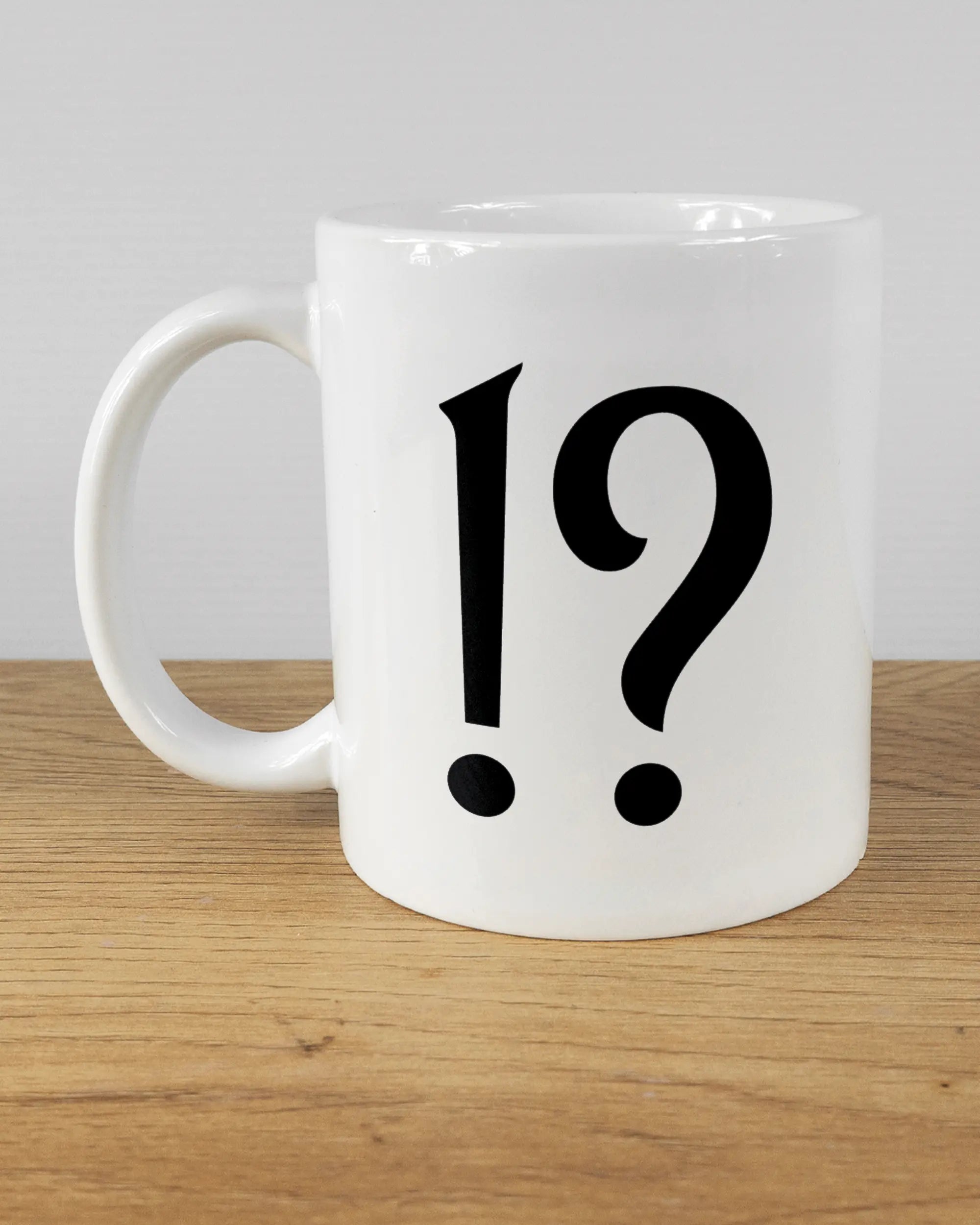 Titelbild, das die linke Seite der beidseitig bedruckten Keramiktasse von Maondo mit dem Design WHAT in der Farbe White auf einem Holztisch zeigt