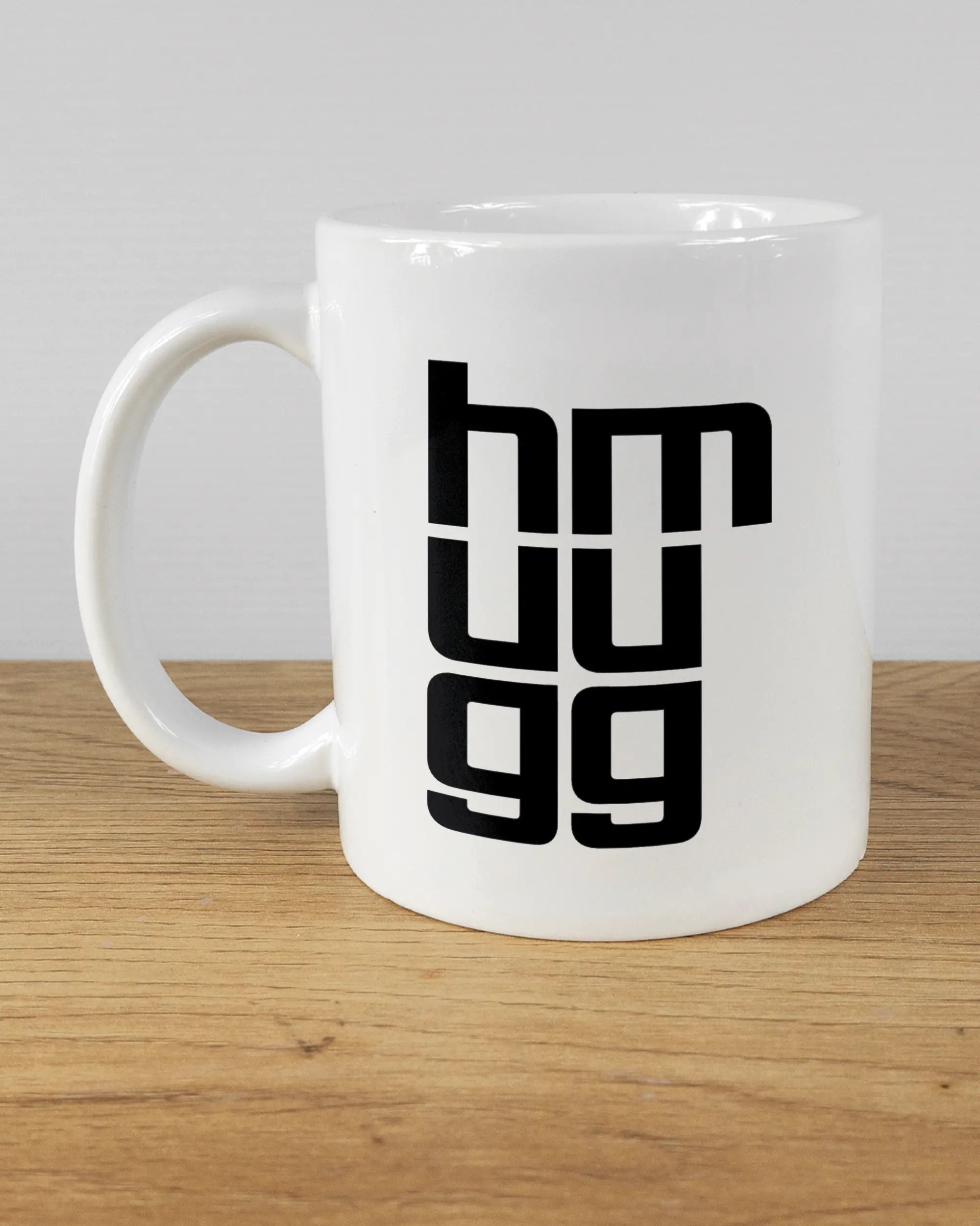 Titelbild, das die linke Seite der beidseitig bedruckten Keramiktasse von Maondo mit dem Design Hug Mug in der Farbe White auf einem Holztisch zeigt