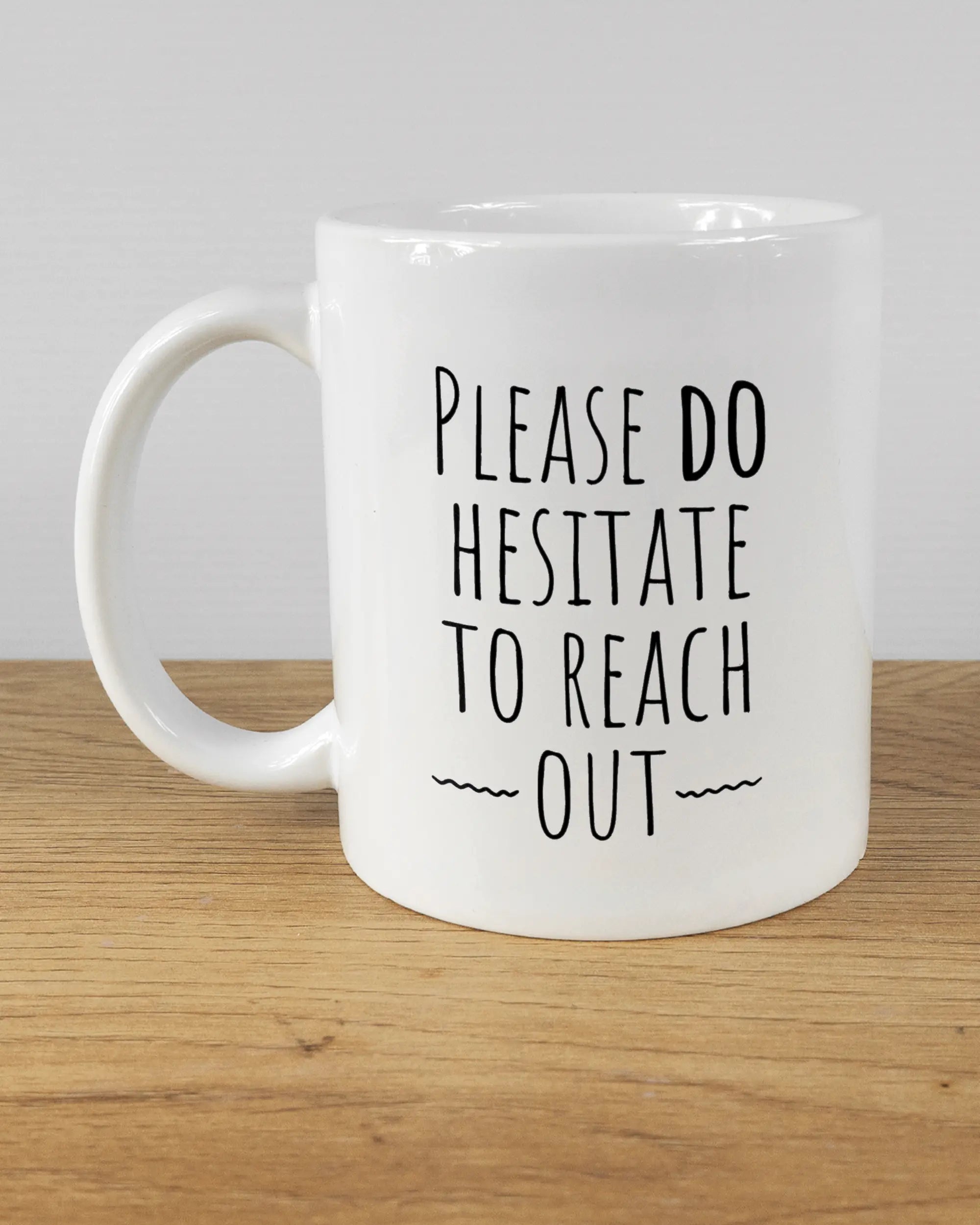 Titelbild, das die linke Seite der beidseitig bedruckten Keramiktasse von Maondo mit dem Design Please Hesitate in der Farbe White auf einem Holztisch zeigt