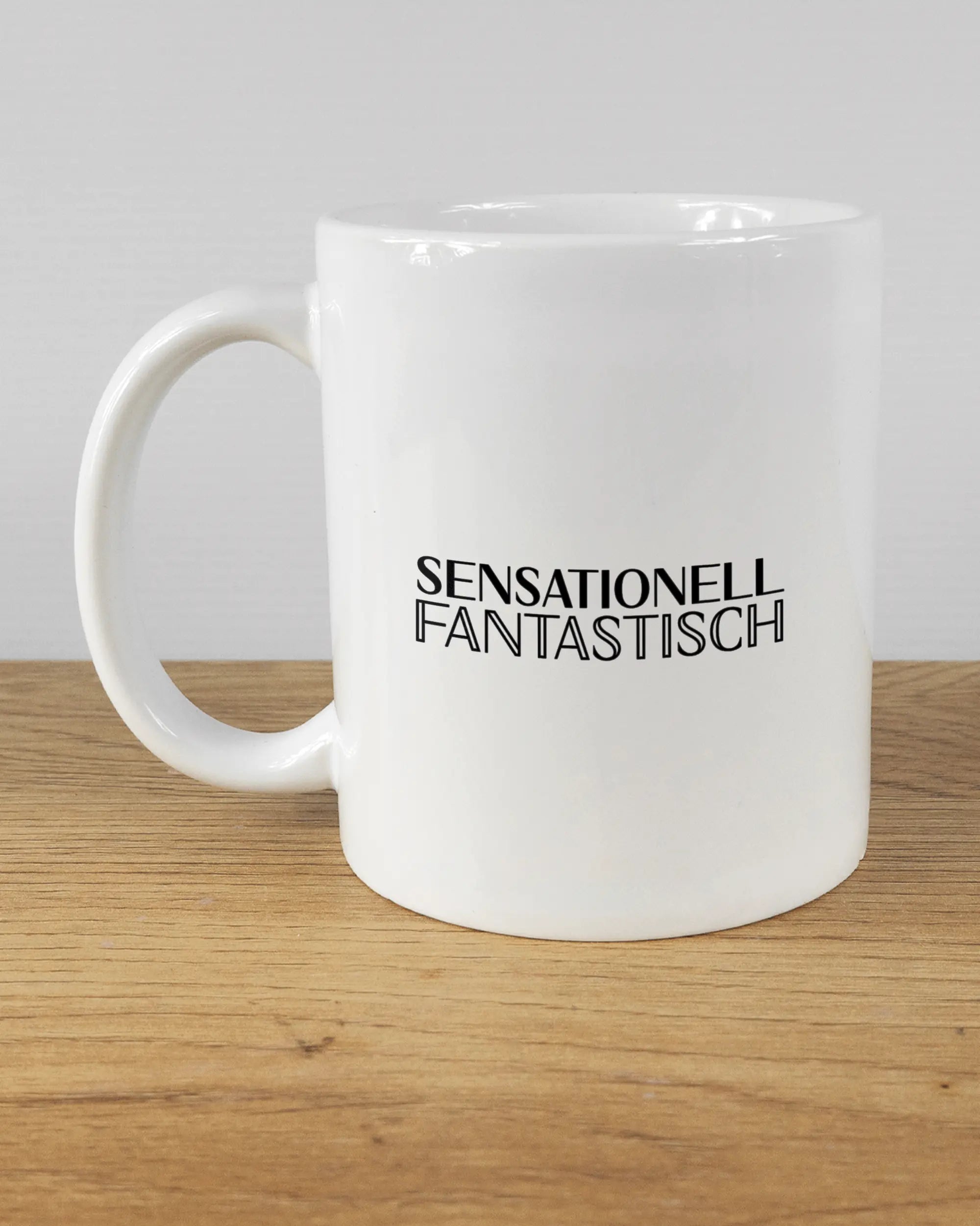 Titelbild, das die linke Seite der beidseitig bedruckten Keramiktasse von Maondo mit dem Design Sensationell in der Farbe White auf einem Holztisch zeigt