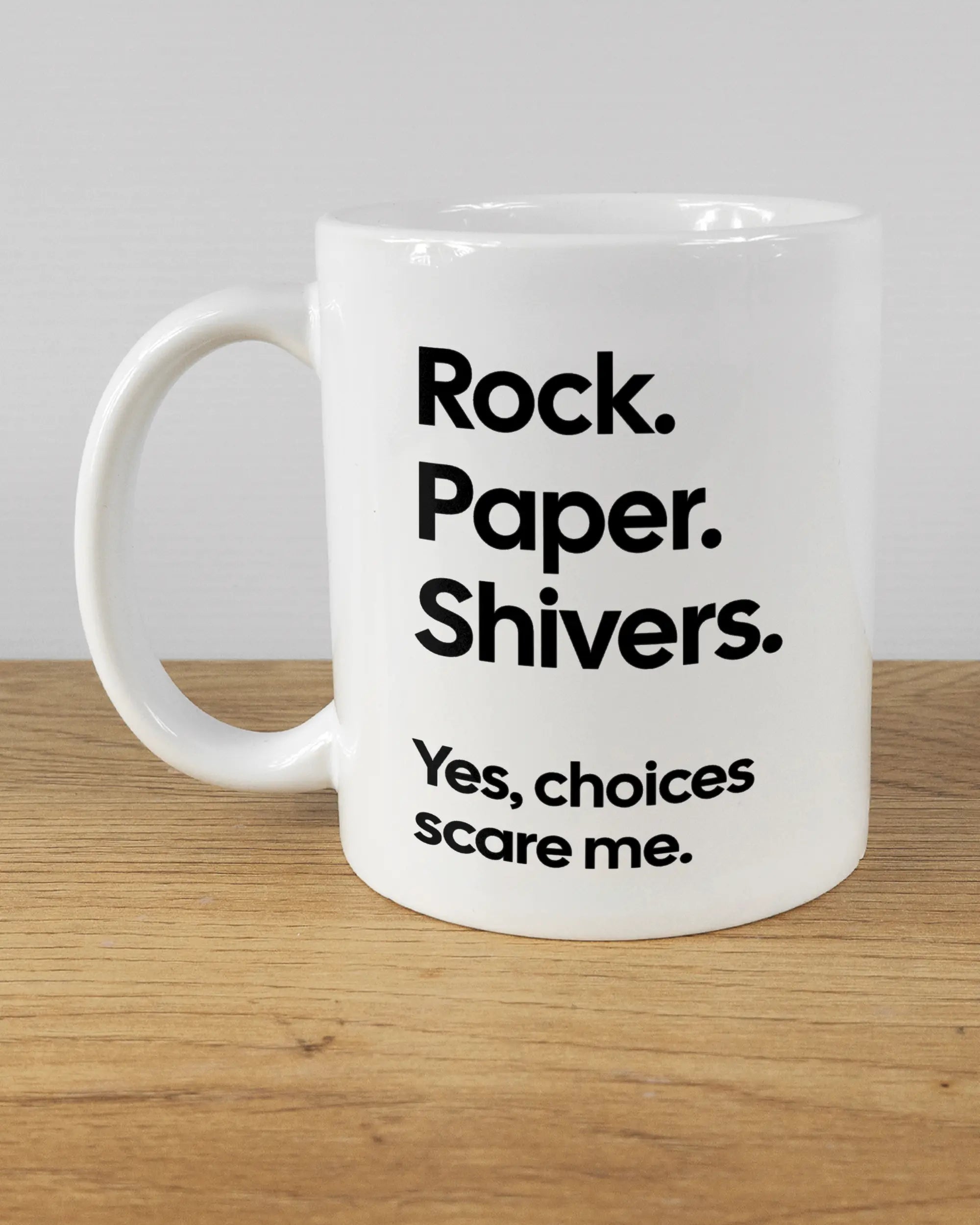 Titelbild, das die linke Seite der beidseitig bedruckten Keramiktasse von Maondo mit dem Design Rock Paper Shivers in der Farbe White auf einem Holztisch zeigt