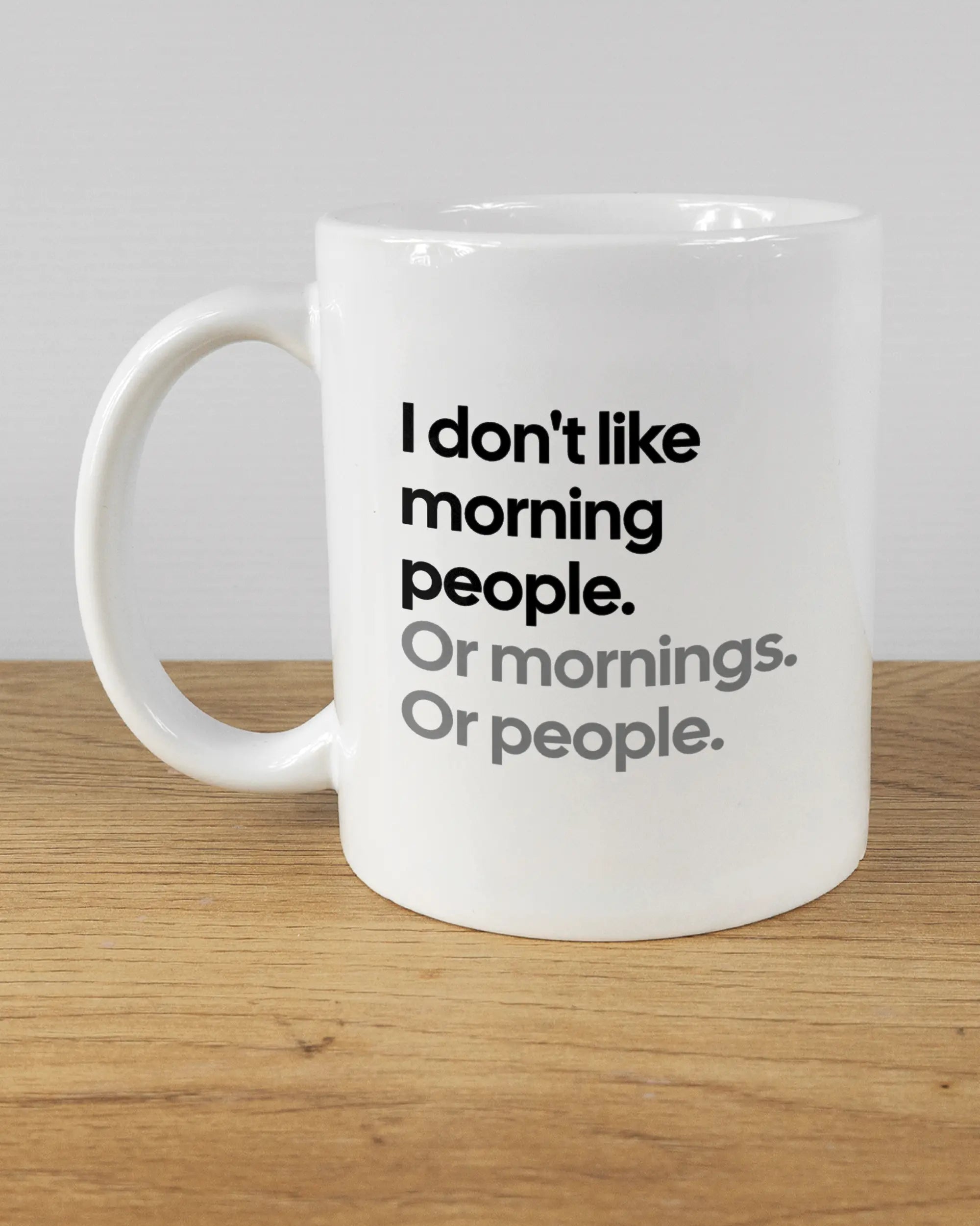 Titelbild, das die linke Seite der beidseitig bedruckten Keramiktasse von Maondo mit dem Design Morning Person in der Farbe White auf einem Holztisch zeigt