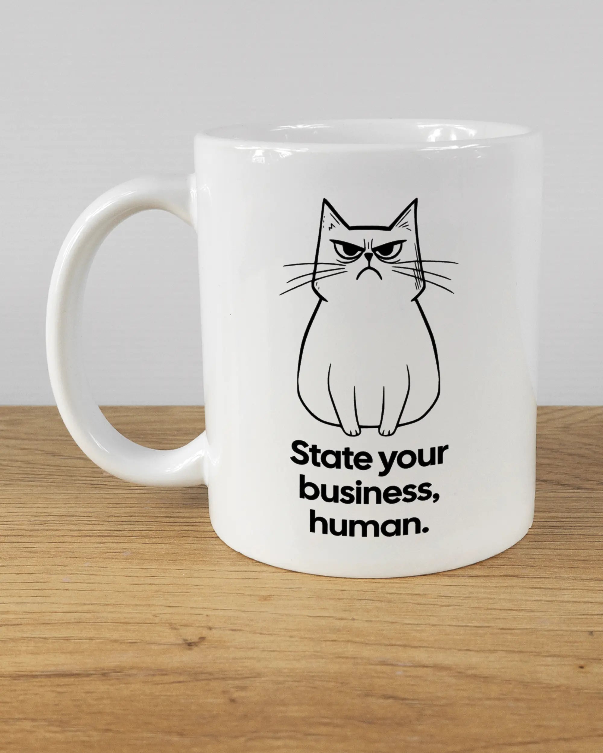 Titelbild, das die linke Seite der beidseitig bedruckten Keramiktasse von Maondo mit dem Design Grumpy Purr in der Farbe White auf einem Holztisch zeigt