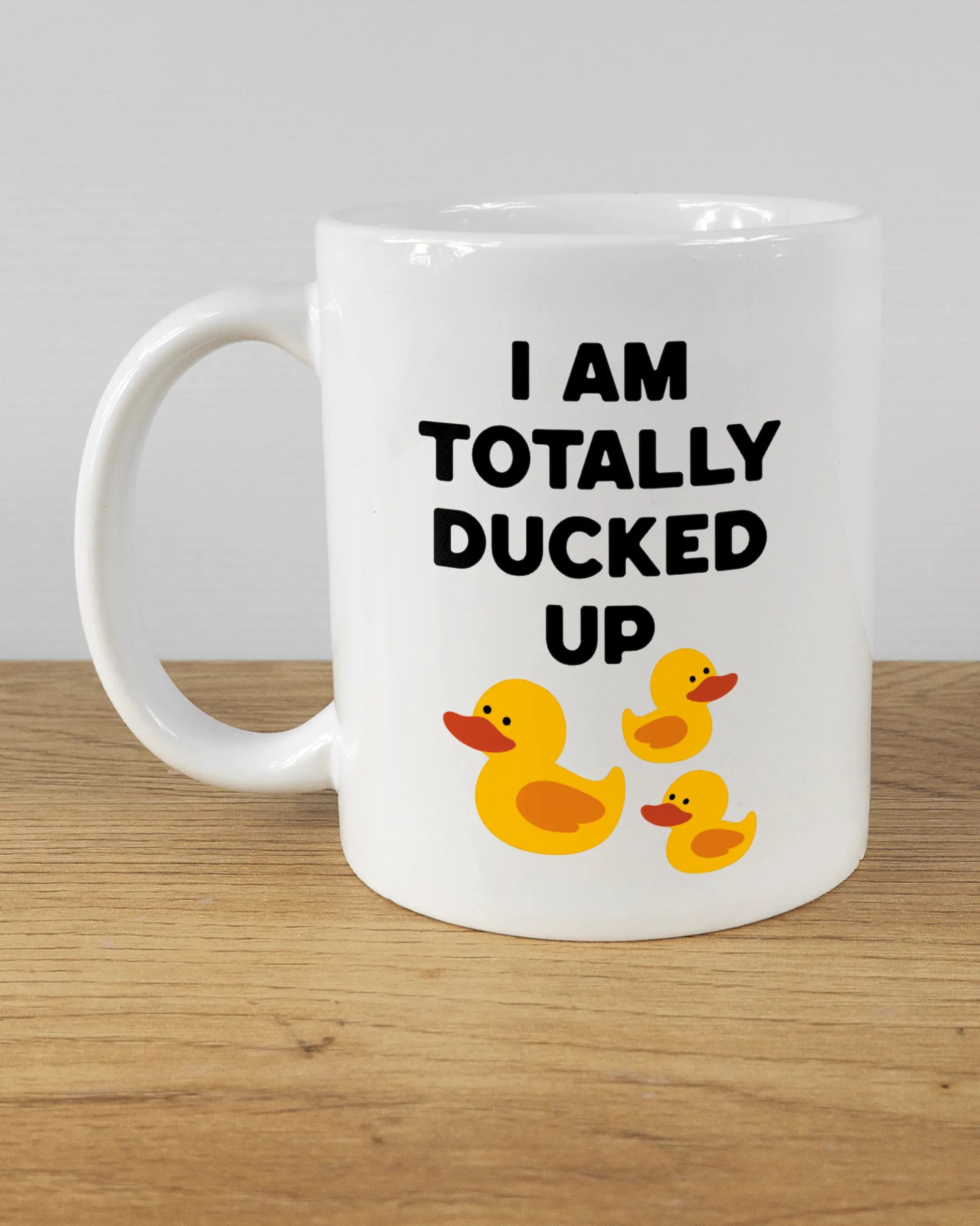 Titelbild, das die linke Seite der beidseitig bedruckten Keramiktasse von Maondo mit dem Design Ducked Up in der Farbe White auf einem Holztisch zeigt