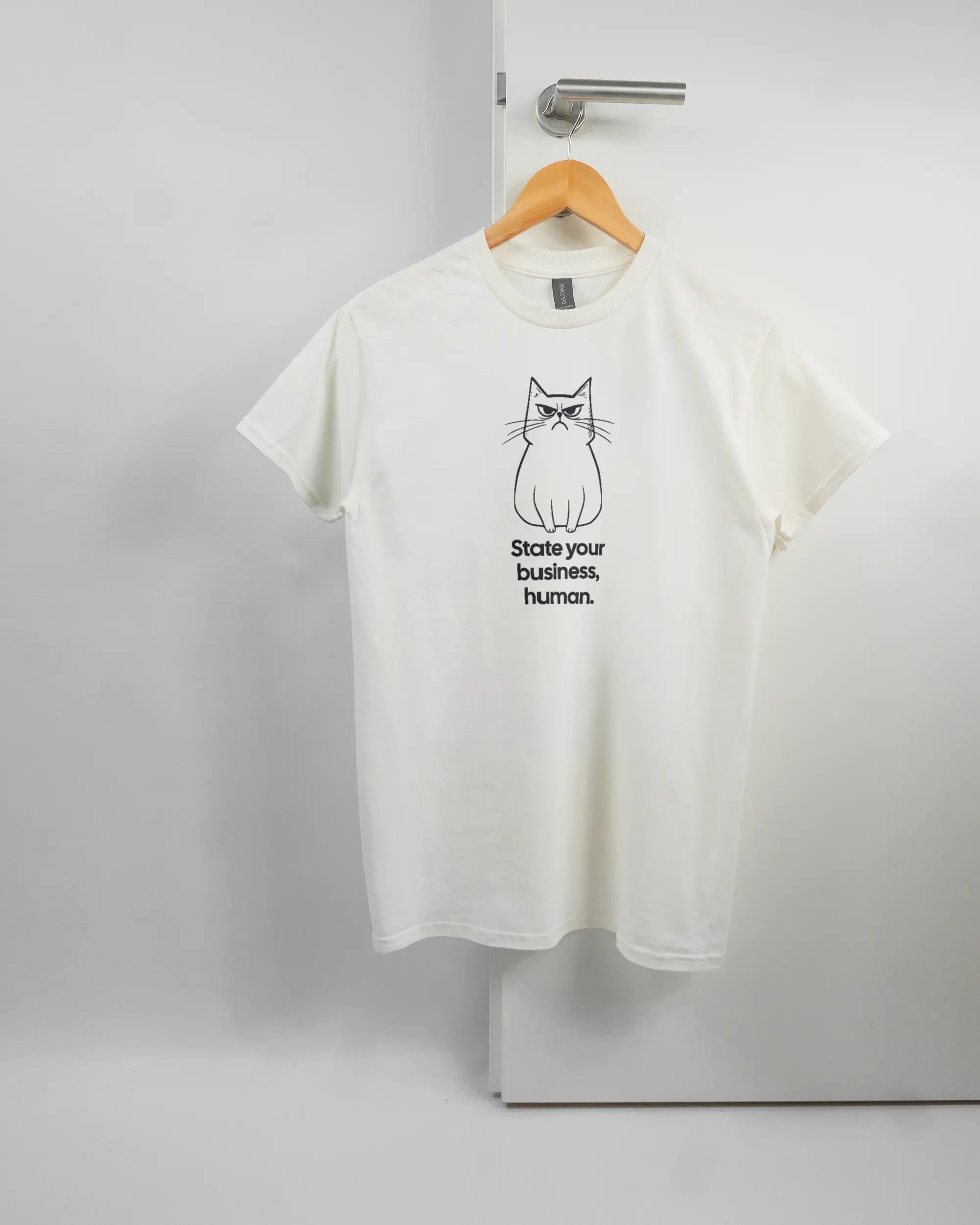 Vorderseite des Basic Unisex T-Shirt Regular von Maondo mit dem Design Grumpy Purr in der Farbe White und in der Größe S auf einem Bügel an der Türklinke aufgehängt