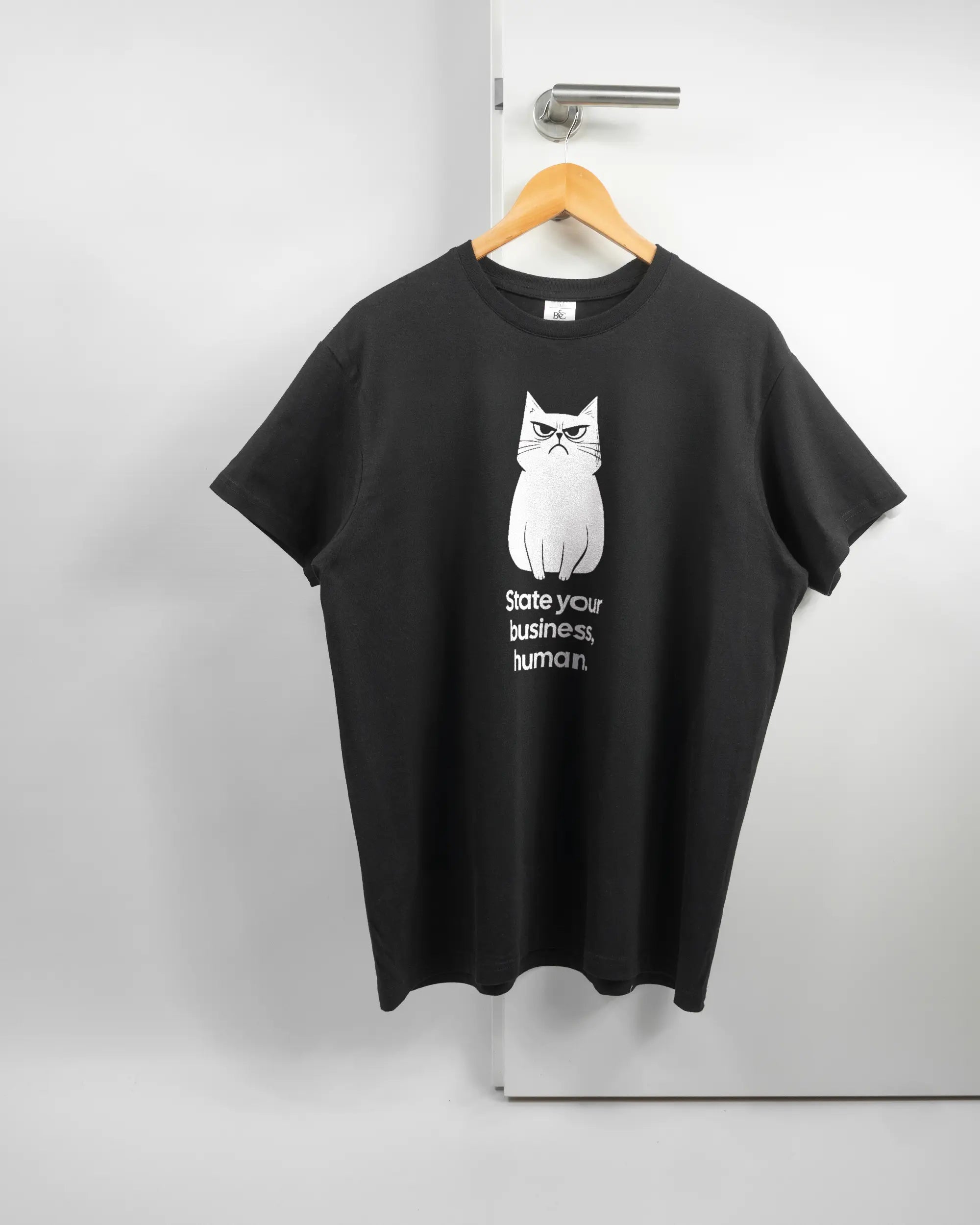 Vorderseite des Basic Unisex T-Shirt Soft von Maondo mit dem Design Grumpy Purr in der Farbe Black und in der Größe L auf einem Bügel an der Türklinke aufgehängt