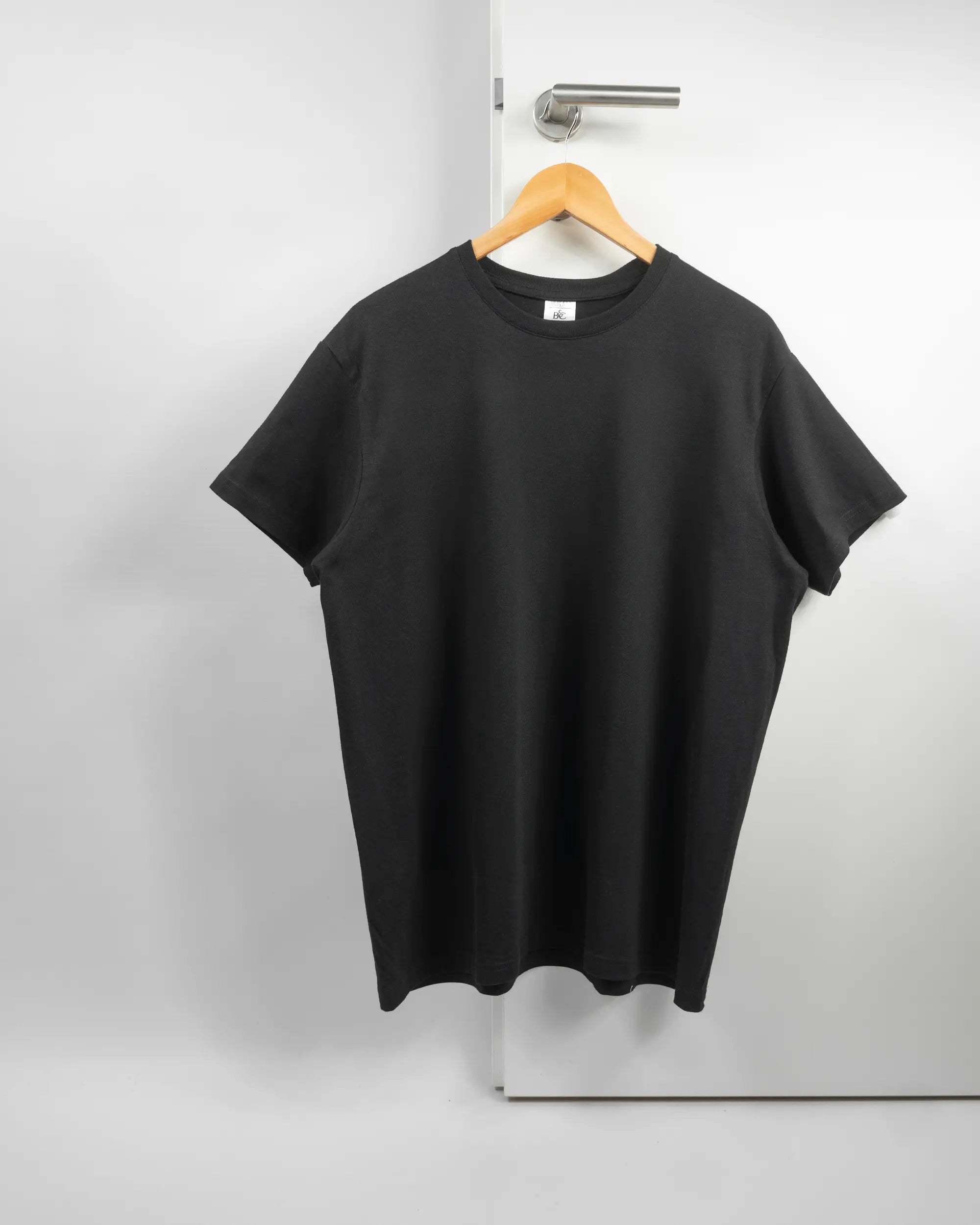 Vorderseite des Basic Unisex T-Shirt Slim von Maondo ohne Motiv in der Farbe Black und in der Größe L auf einem Bügel an der Türklinke aufgehängt