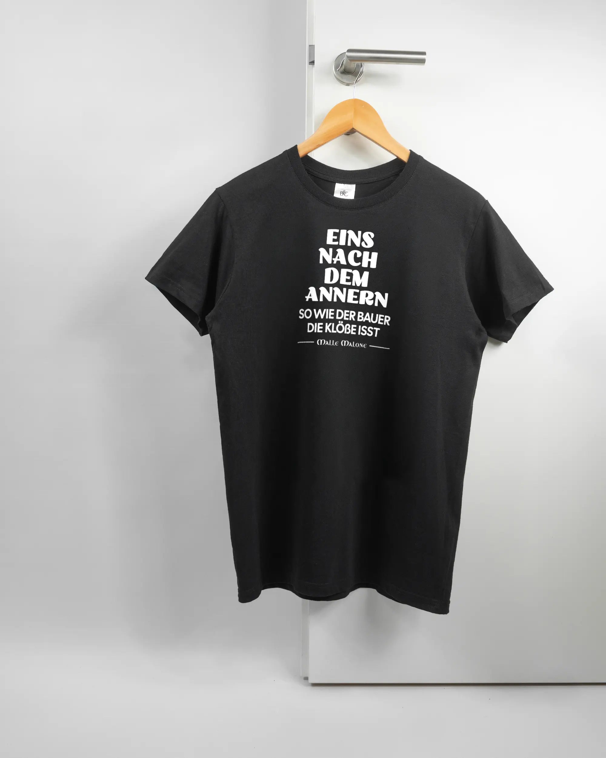 Vorderseite des Basic Unisex T-Shirt Soft von Maondo mit dem Design Eins nach dem Annern in der Farbe Black und in der Größe S auf einem Bügel an der Türklinke aufgehängt