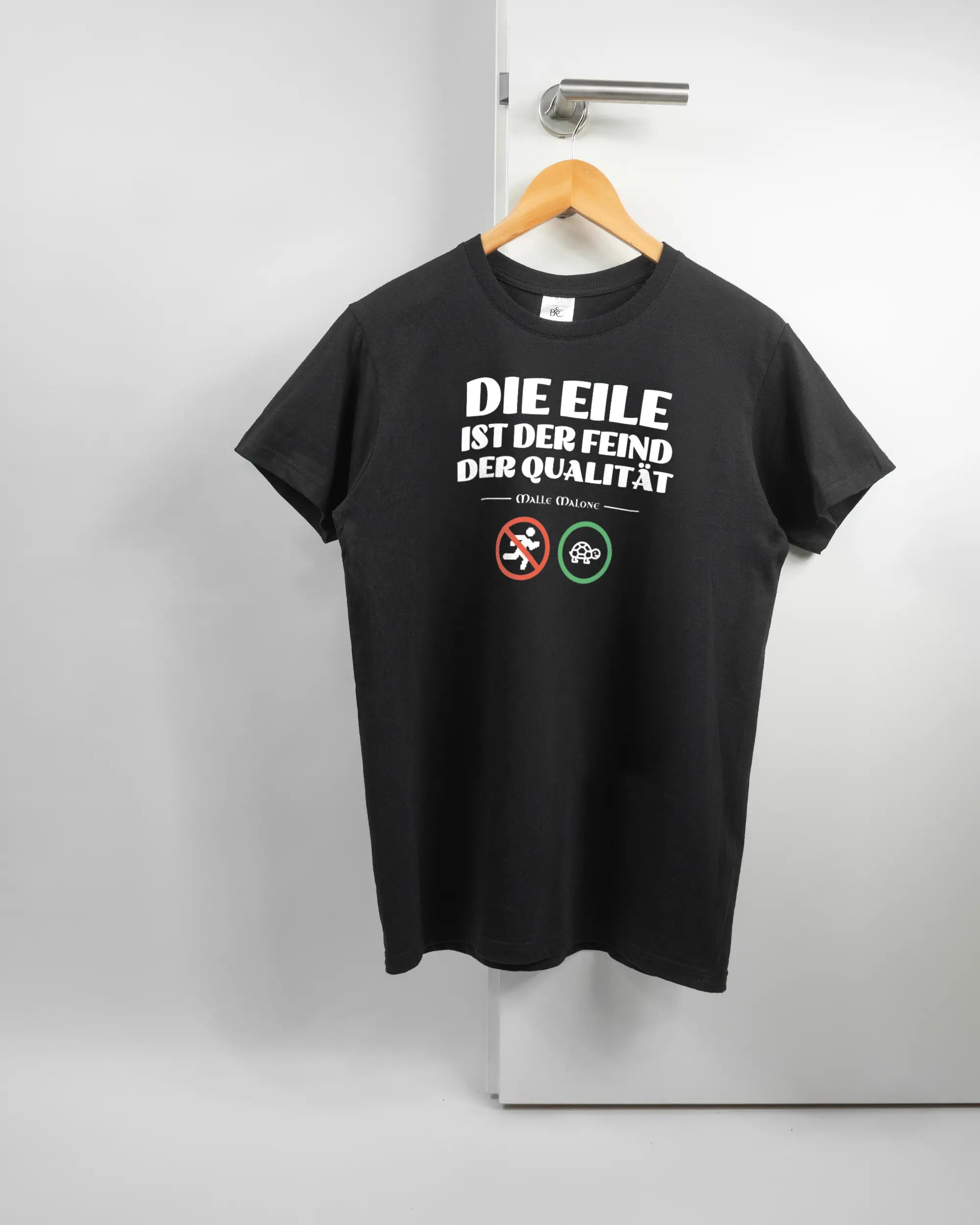 Vorderseite des Basic Unisex T-Shirt Soft von Maondo mit dem Design Keine Eile in der Farbe Black und in der Größe S auf einem Bügel an der Türklinke aufgehängt