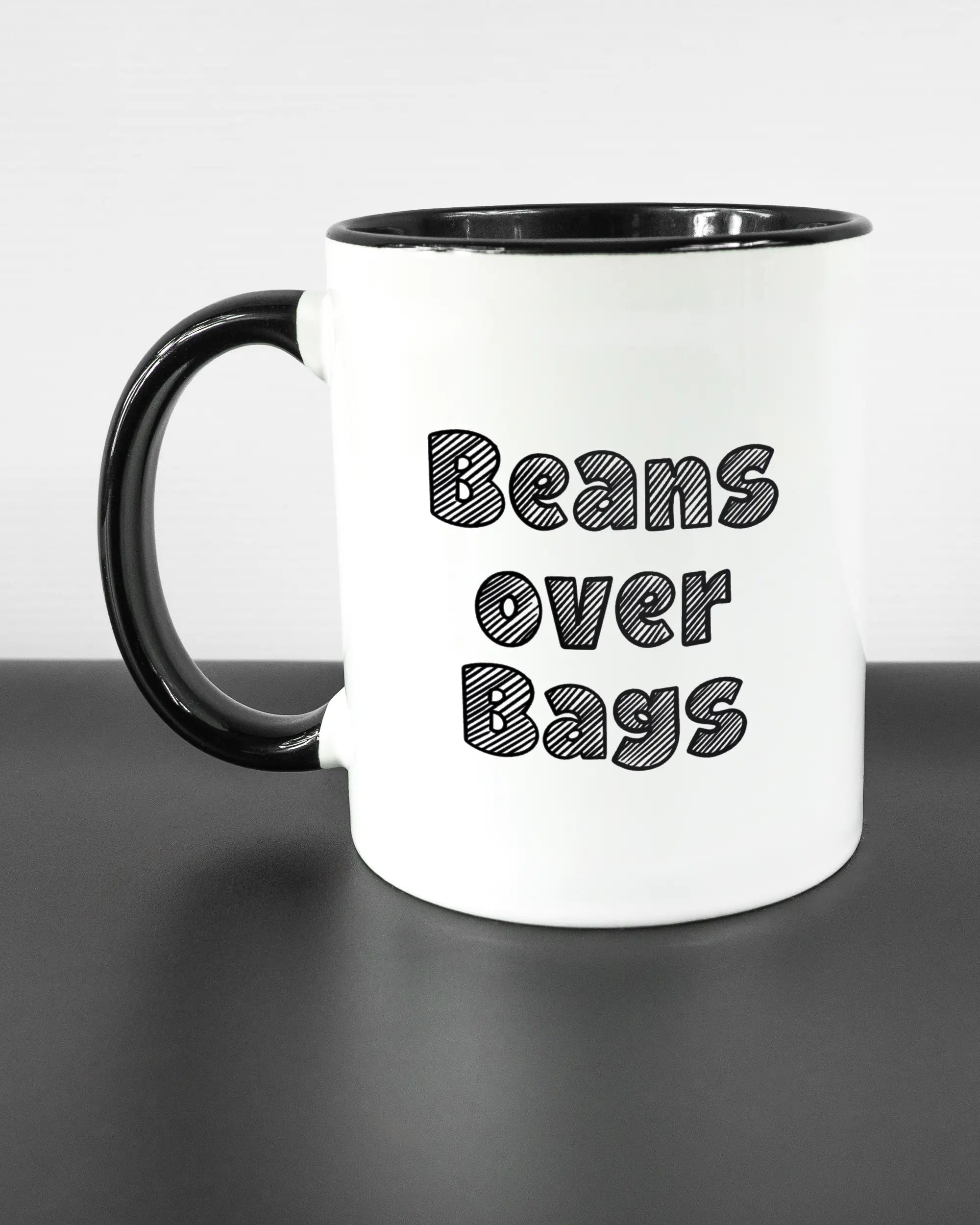 Titelbild, das die linke Seite der beidseitig bedruckten Keramiktasse von Maondo mit dem Design Beans over Bags in der Farbe Duotone auf einem Holztisch zeigt