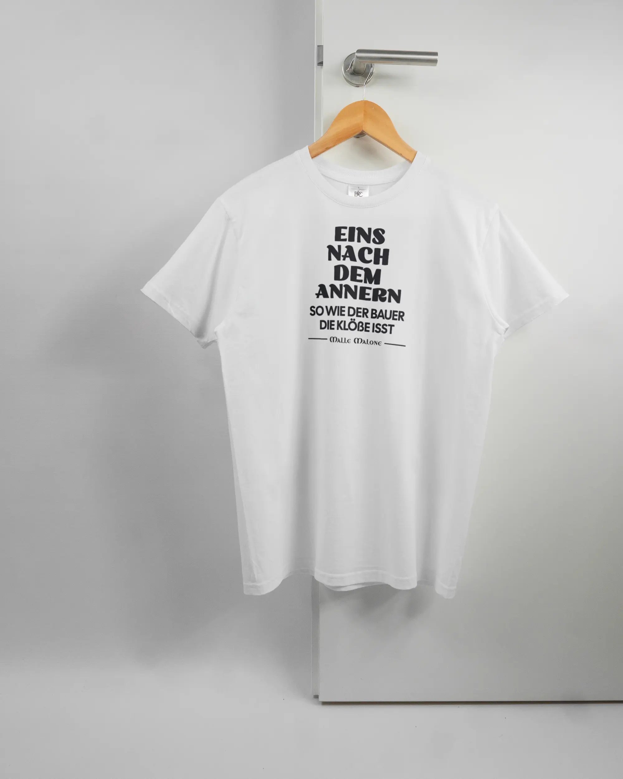 Vorderseite des Basic Unisex T-Shirt Soft von Maondo mit dem Design Eins nach dem Annern in der Farbe White und in der Größe S auf einem Bügel an der Türklinke aufgehängt