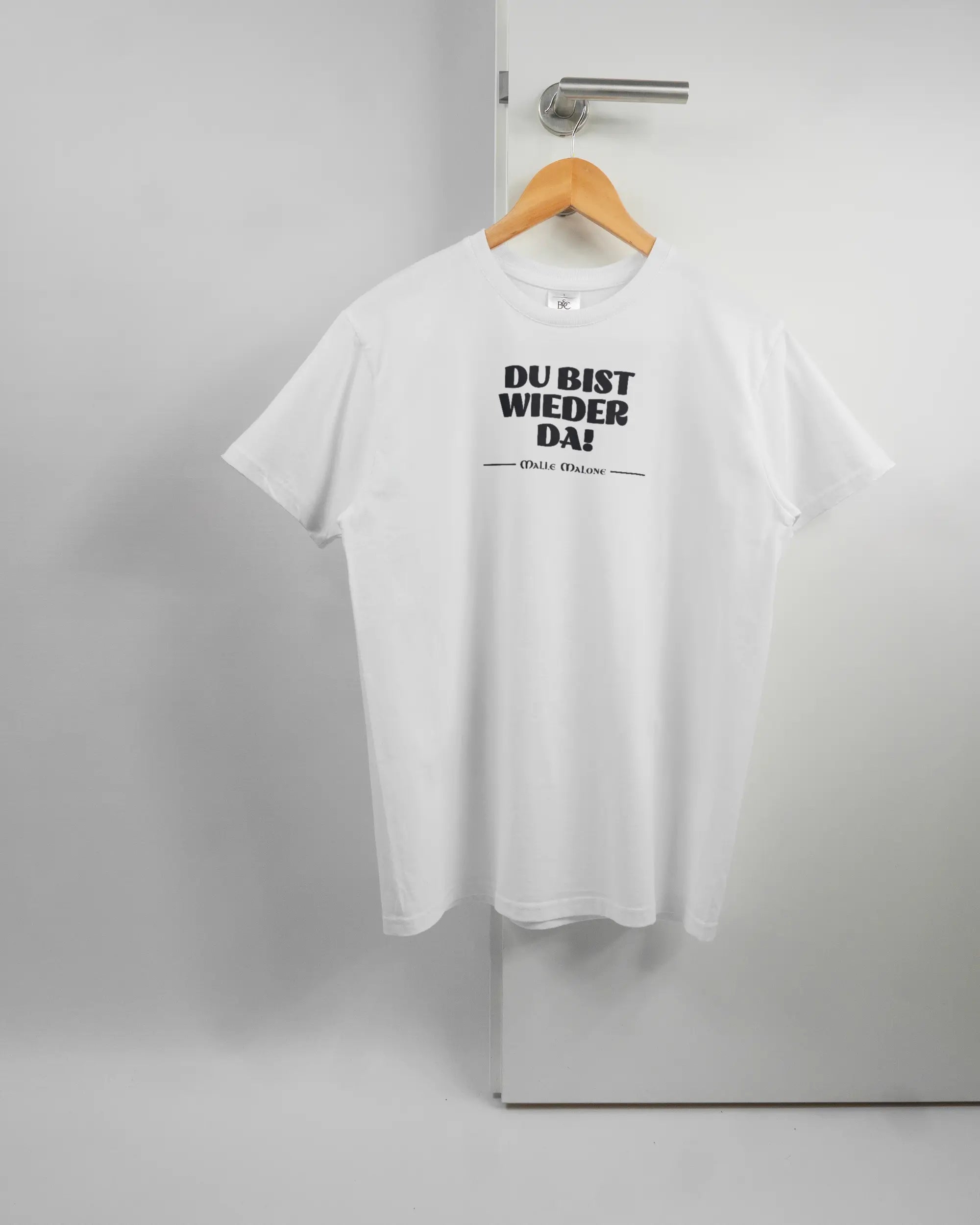 Vorderseite des Basic Unisex T-Shirt Soft von Maondo mit dem Design Wieder da in der Farbe White und in der Größe S auf einem Bügel an der Türklinke aufgehängt