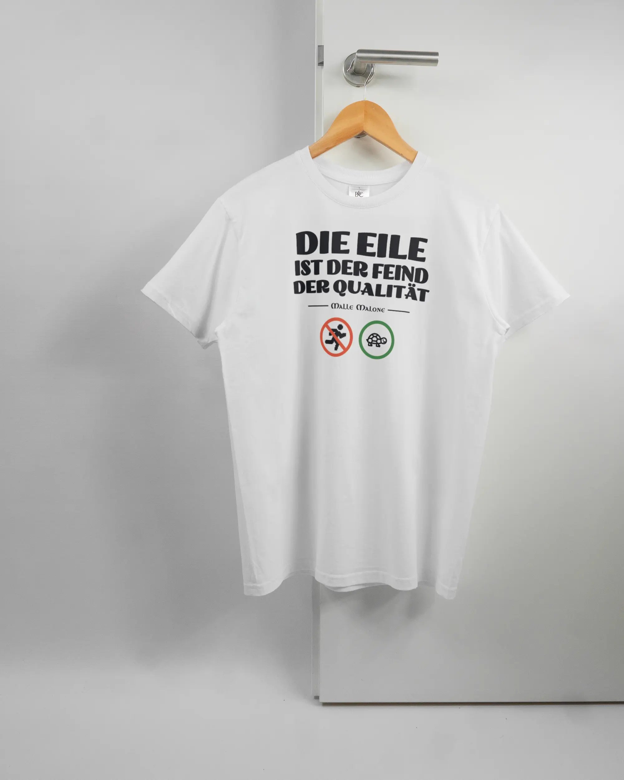 Vorderseite des Basic Unisex T-Shirt Soft von Maondo mit dem Design Keine Eile in der Farbe White und in der Größe S auf einem Bügel an der Türklinke aufgehängt