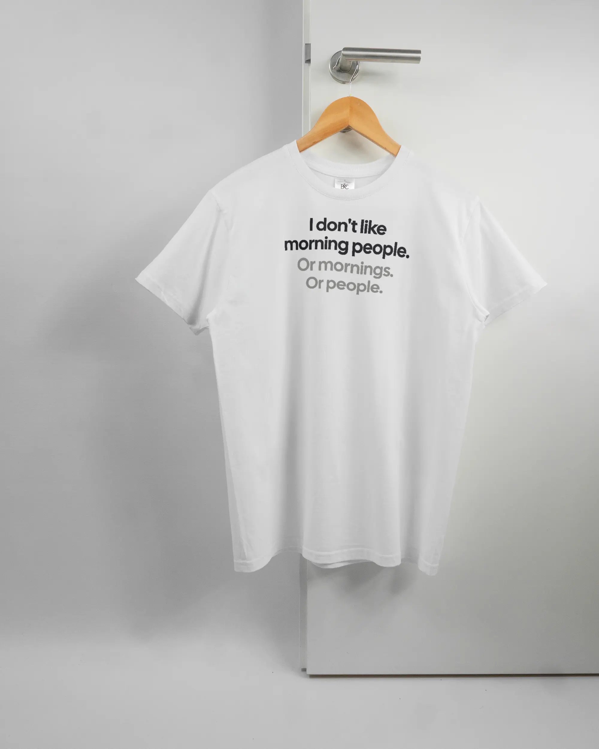 Vorderseite des Basic Unisex T-Shirt Soft von Maondo mit dem Design Morning Person in der Farbe White und in der Größe S auf einem Bügel an der Türklinke aufgehängt