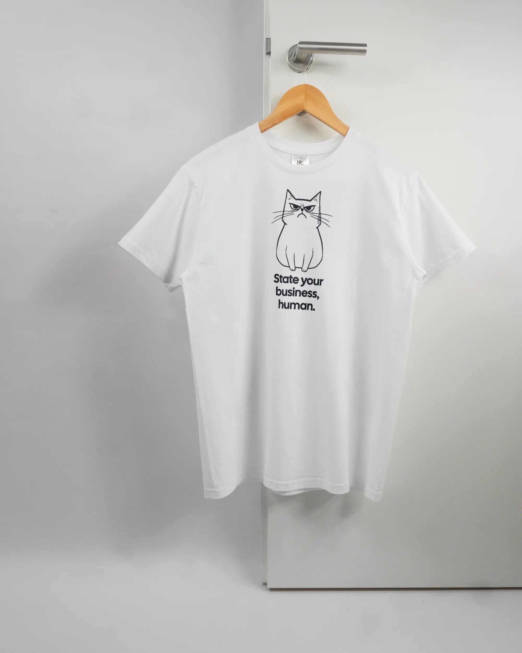 Vorderseite des Basic Unisex T-Shirt Soft von Maondo mit dem Design Grumpy Purr in der Farbe White und in der Größe S auf einem Bügel an der Türklinke aufgehängt
