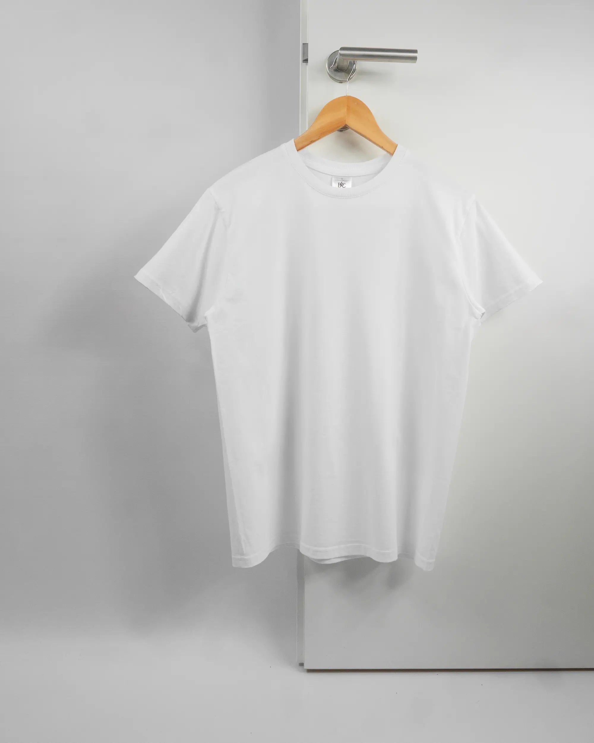 Vorderseite des Basic Unisex T-Shirt Slim von Maondo ohne Motiv in der Farbe White und in der Größe S auf einem Bügel an der Türklinke aufgehängt