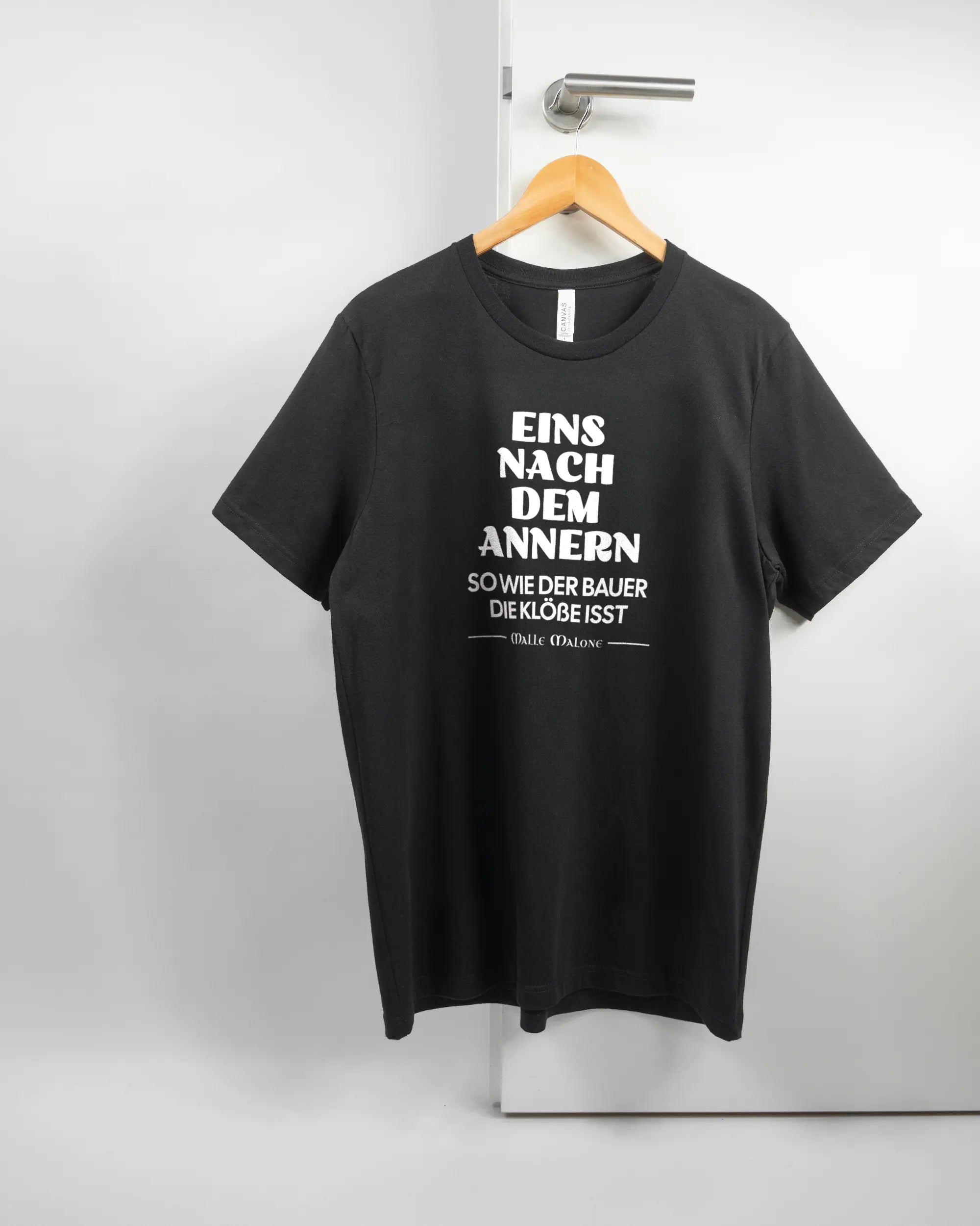 Vorderseite des Basic Unisex T-Shirt Light von Maondo mit dem Design Eins nach dem Annern in der Farbe Black und in der Größe L auf einem Bügel an der Türklinke aufgehängt