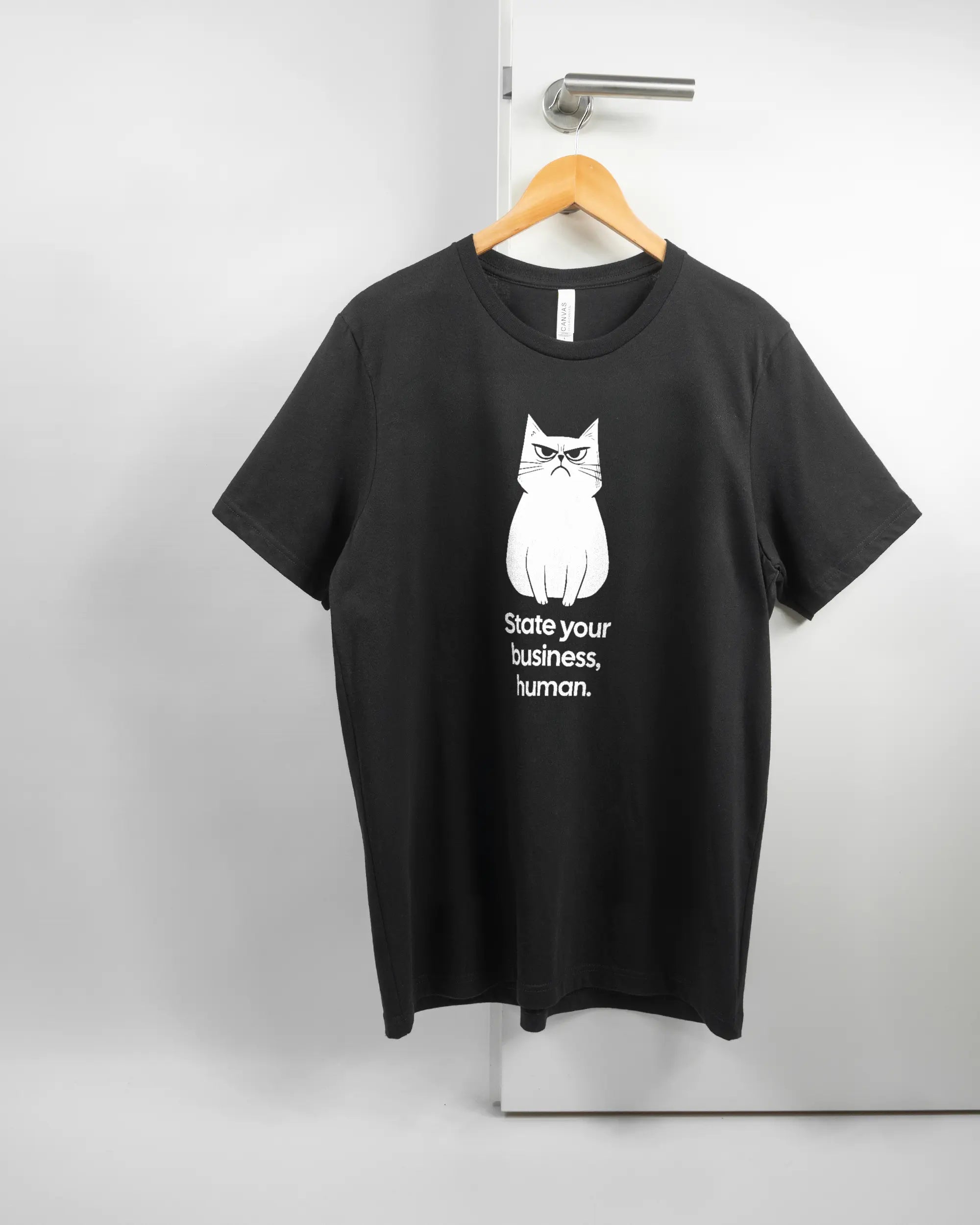 Vorderseite des Basic Unisex T-Shirt Light von Maondo mit dem Design Grumpy Purr in der Farbe Black und in der Größe L auf einem Bügel an der Türklinke aufgehängt