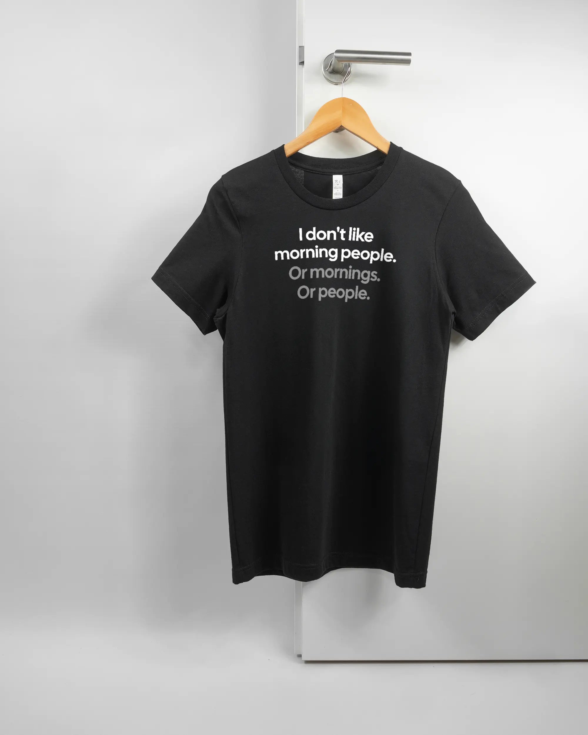 Vorderseite des Basic Unisex T-Shirt Light von Maondo mit dem Design Morning Person in der Farbe Black und in der Größe S auf einem Bügel an der Türklinke aufgehängt