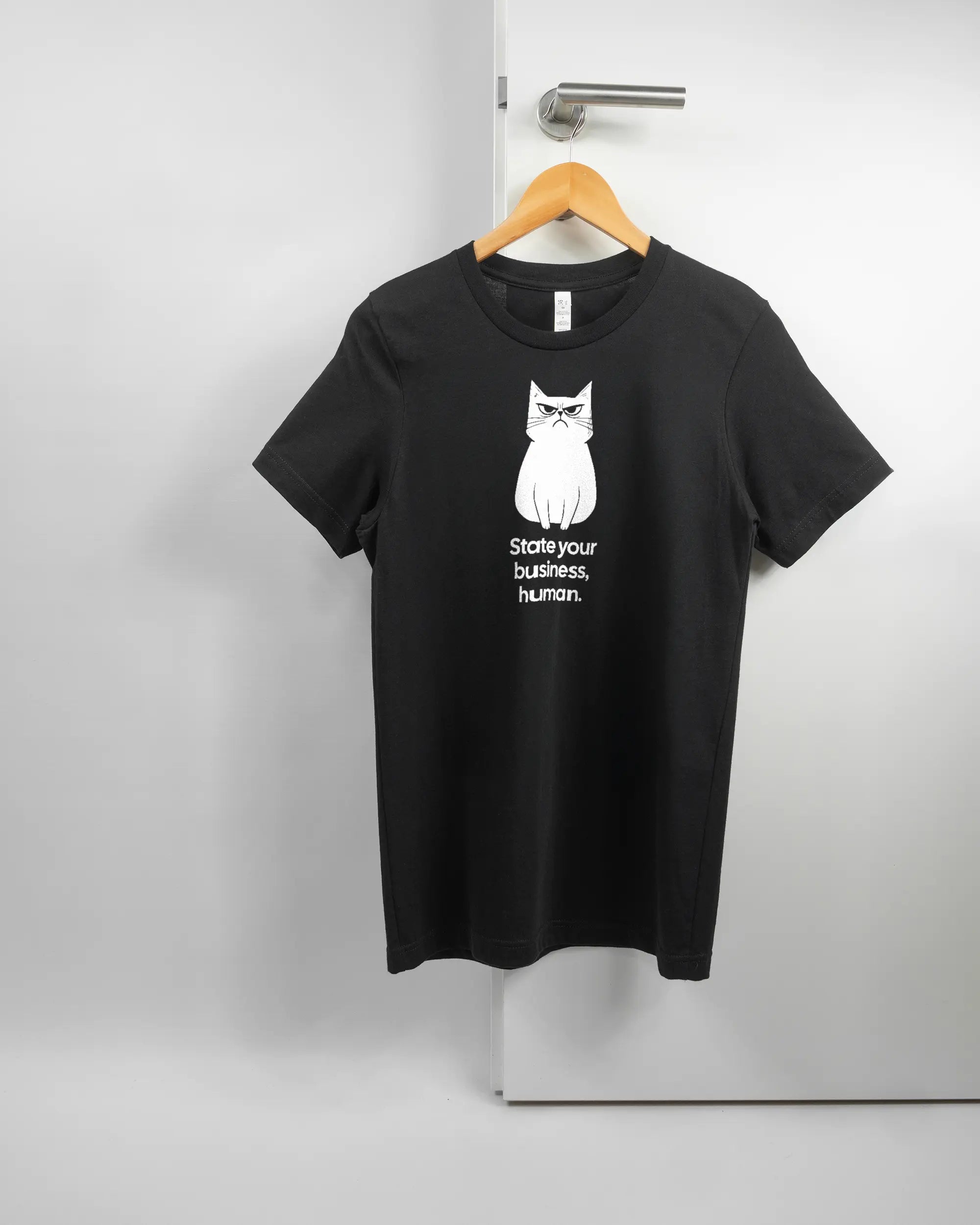 Vorderseite des Basic Unisex T-Shirt Light von Maondo mit dem Design Grumpy Purr in der Farbe Black und in der Größe S auf einem Bügel an der Türklinke aufgehängt