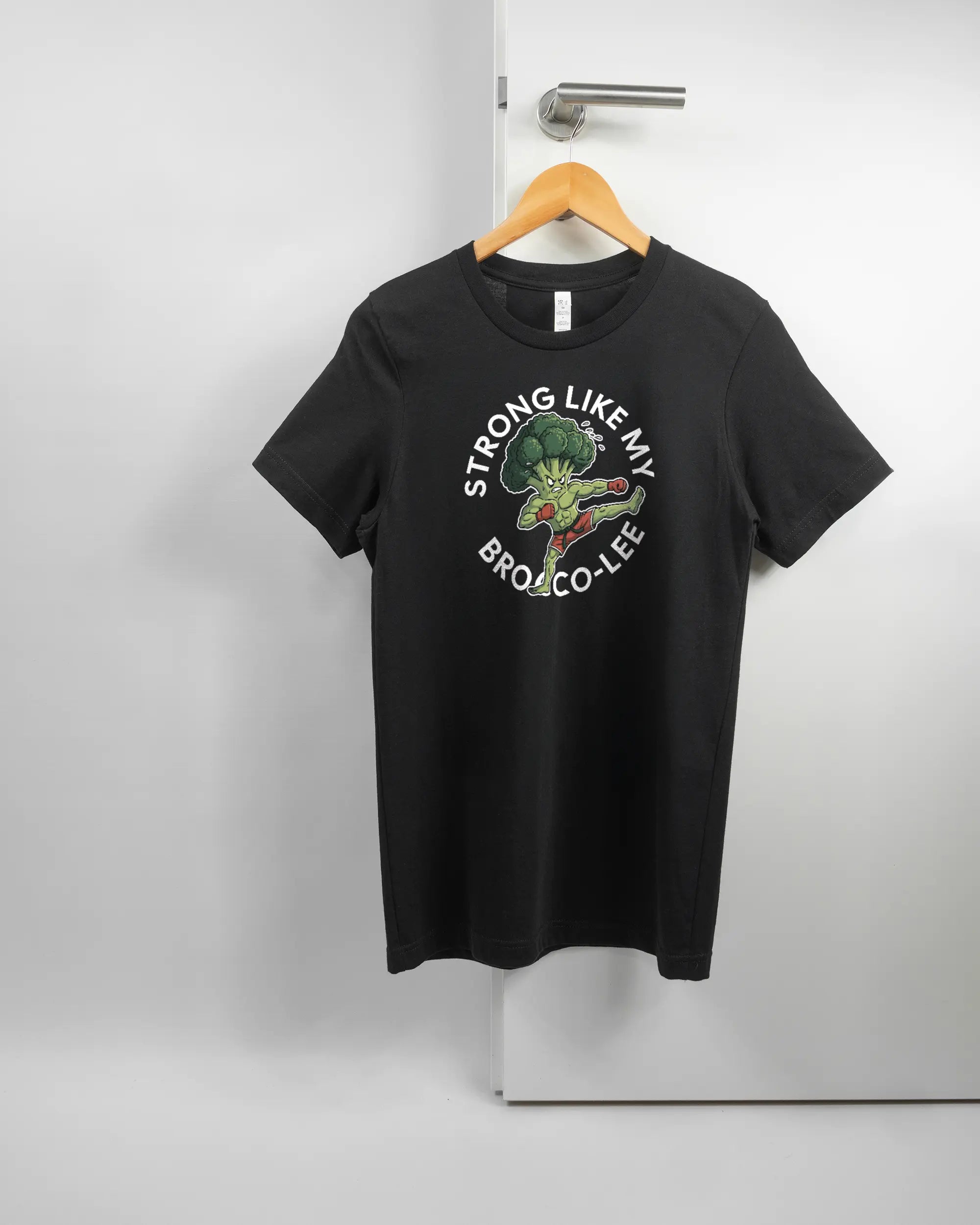 Vorderseite des Basic Unisex T-Shirt Light von Maondo mit dem Design Brocco-Lee in der Farbe Black und in der Größe S auf einem Bügel an der Türklinke aufgehängt