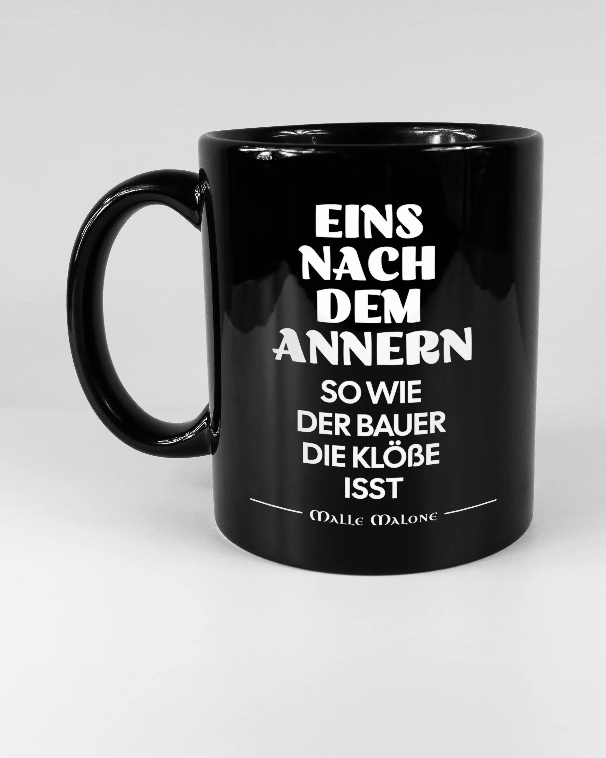 Titelbild, das die linke Seite der beidseitig bedruckten Keramiktasse von Maondo mit dem Design Eins nach dem Annern in der Farbe Black auf einem Holztisch zeigt