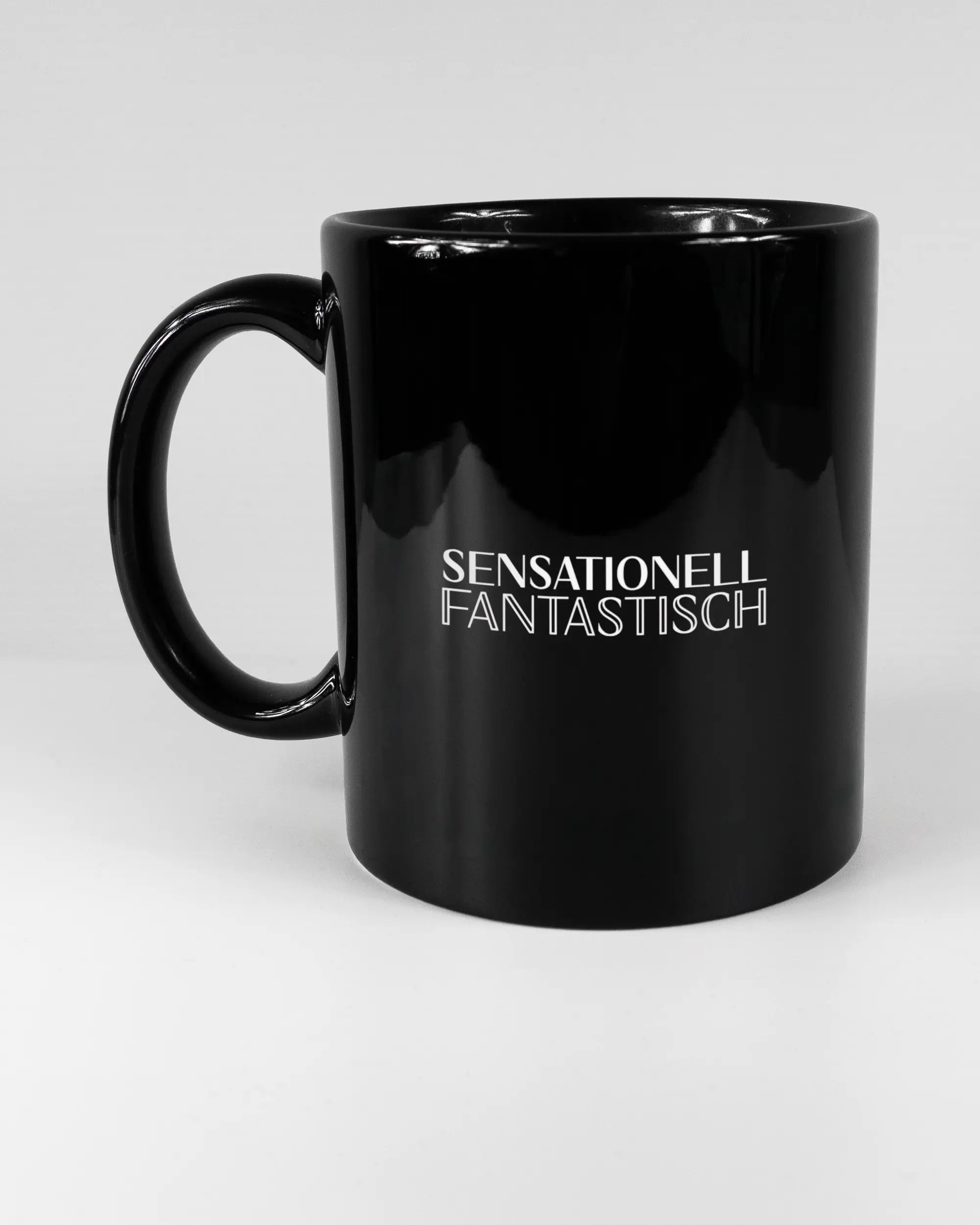 Titelbild, das die linke Seite der beidseitig bedruckten Keramiktasse von Maondo mit dem Design Sensationell in der Farbe Black auf einem Holztisch zeigt