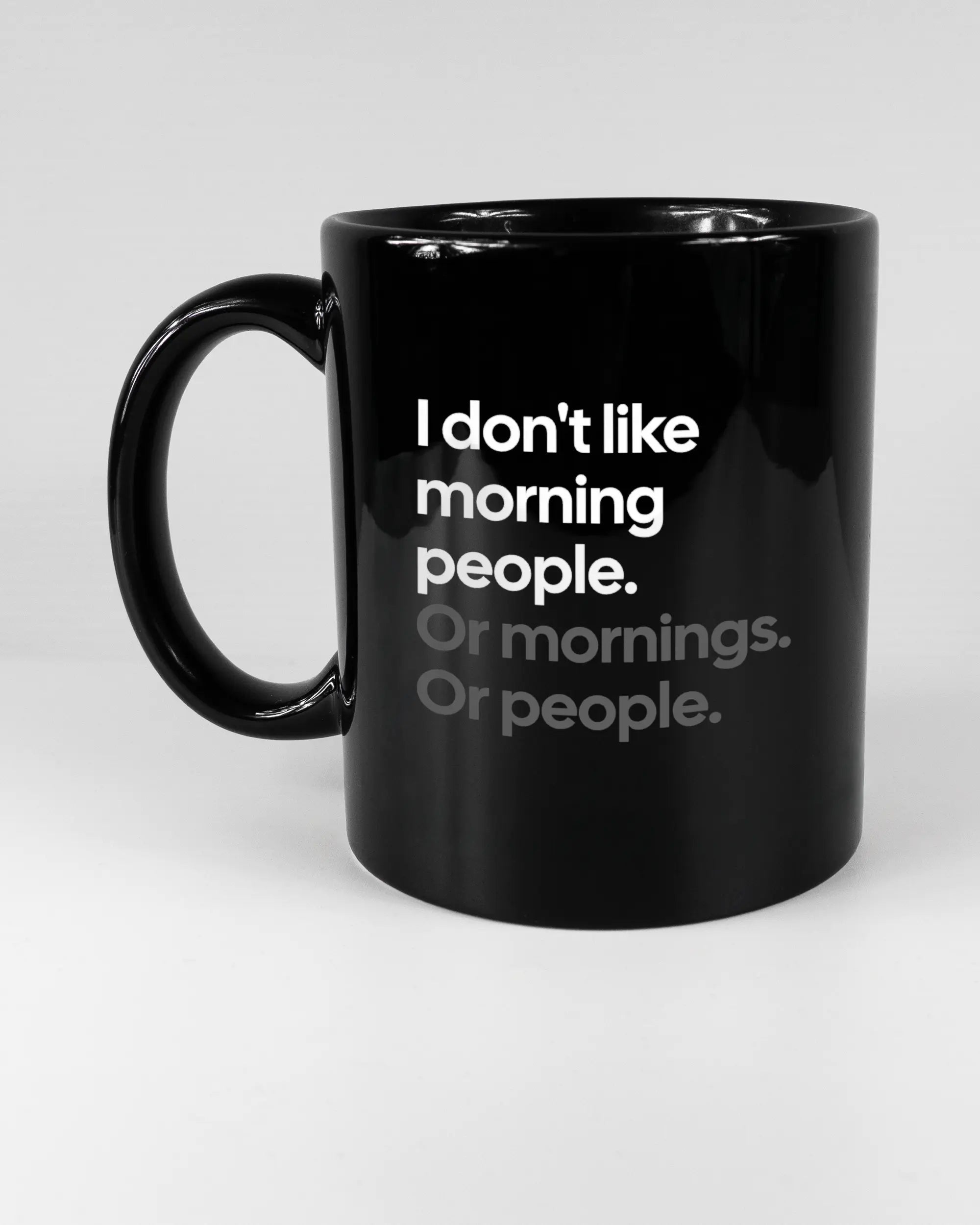 Titelbild, das die linke Seite der beidseitig bedruckten Keramiktasse von Maondo mit dem Design Morning Person in der Farbe Black auf einem Holztisch zeigt