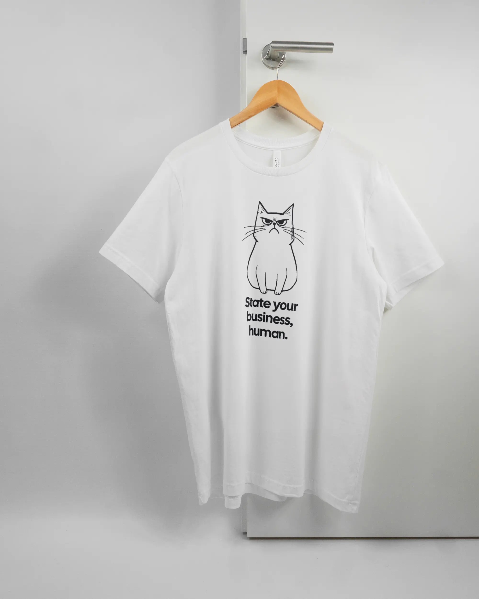 Vorderseite des Basic Unisex T-Shirt Light von Maondo mit dem Design Grumpy Purr in der Farbe White und in der Größe L auf einem Bügel an der Türklinke aufgehängt