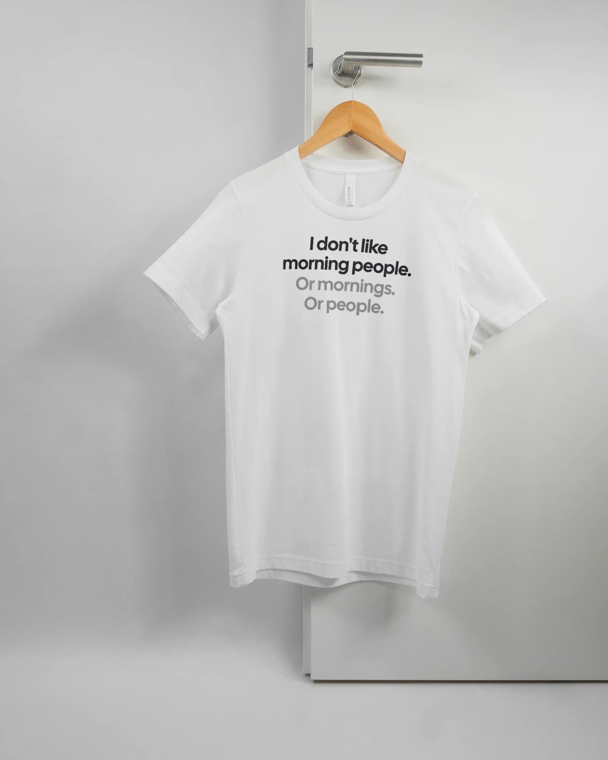 Vorderseite des Basic Unisex T-Shirt Light von Maondo mit dem Design Morning Person in der Farbe White und in der Größe S auf einem Bügel an der Türklinke aufgehängt