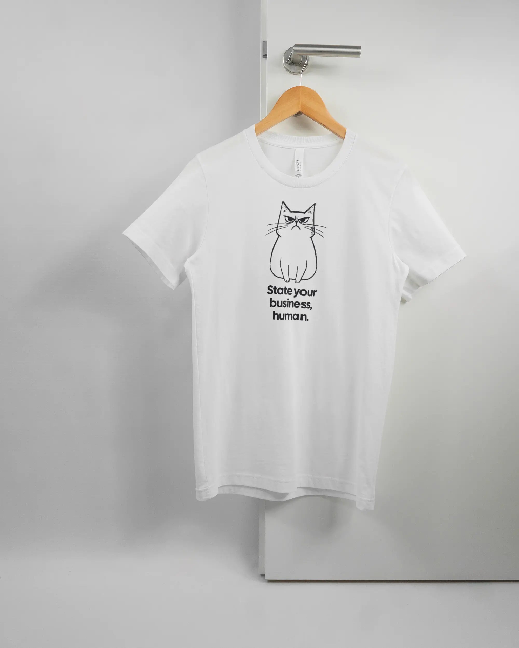 Vorderseite des Basic Unisex T-Shirt Light von Maondo mit dem Design Grumpy Purr in der Farbe White und in der Größe S auf einem Bügel an der Türklinke aufgehängt