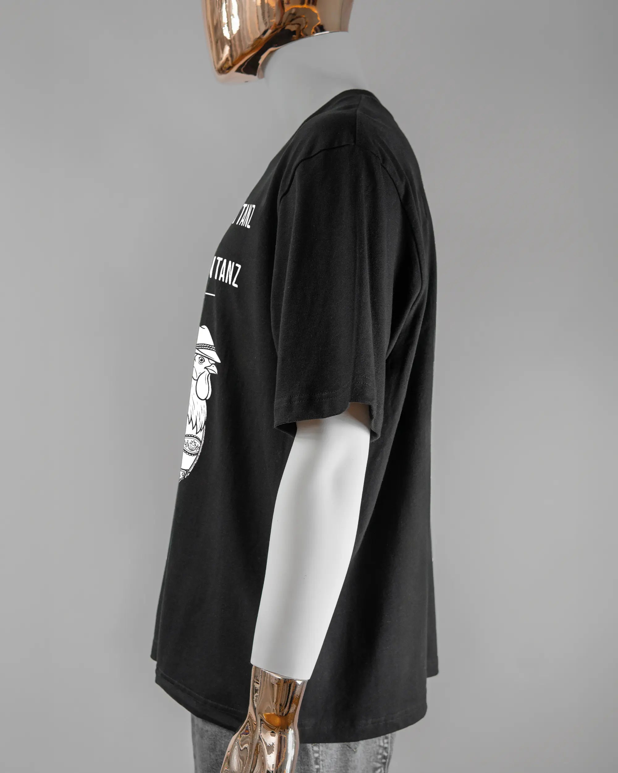 Seitenansicht des Basic Unisex T-Shirt Oversized von Maondo mit dem Design Trachtentanz vorne in der Farbe Black und in der Größe L getragen von einer maskulinen Puppe