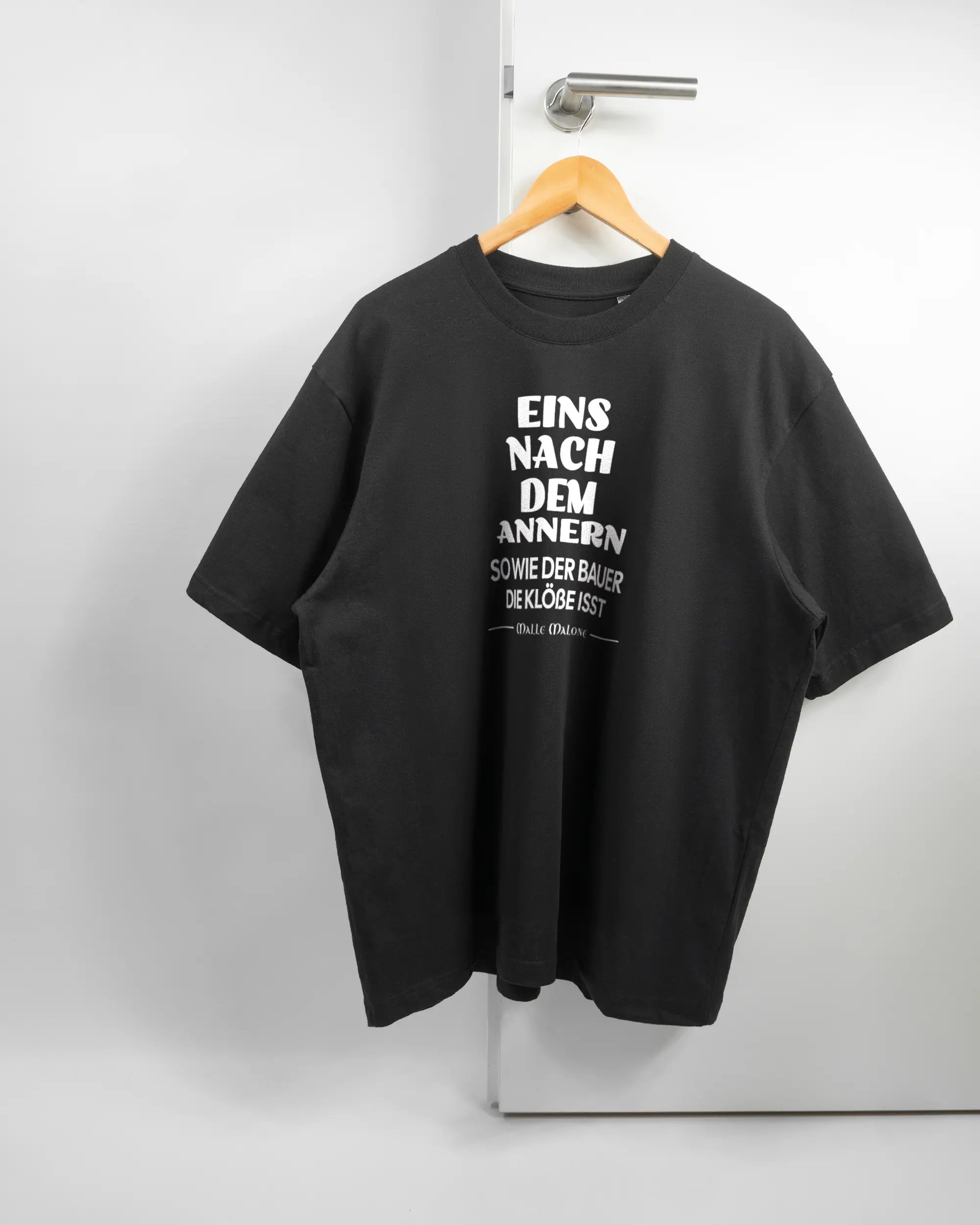 Vorderseite des Basic Unisex T-Shirt Oversized von Maondo mit dem Design Eins nach dem Annern in der Farbe Black und in der Größe L auf einem Bügel an der Türklinke aufgehängt