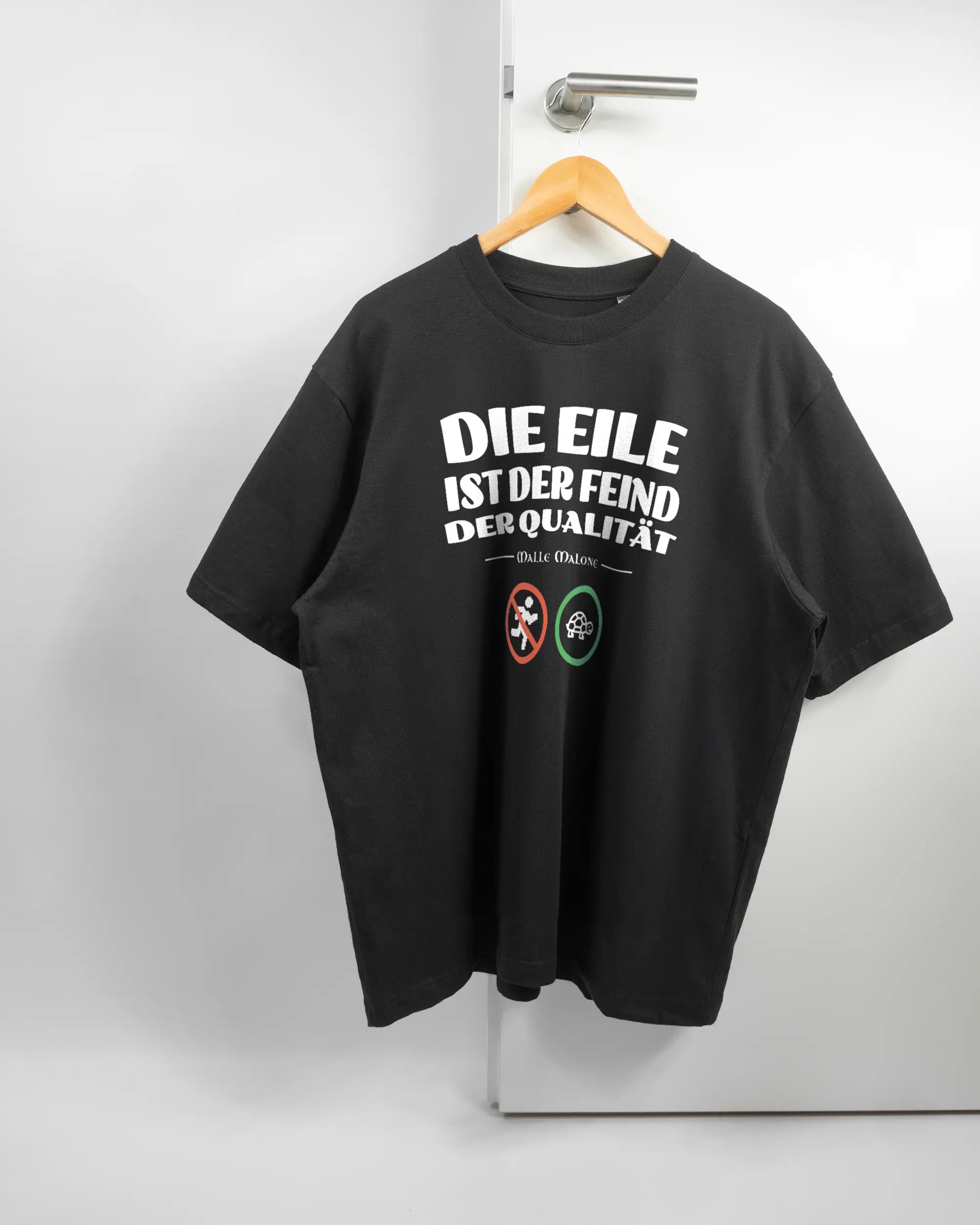 Vorderseite des Basic Unisex T-Shirt Oversized von Maondo mit dem Design Keine Eile in der Farbe Black und in der Größe L auf einem Bügel an der Türklinke aufgehängt