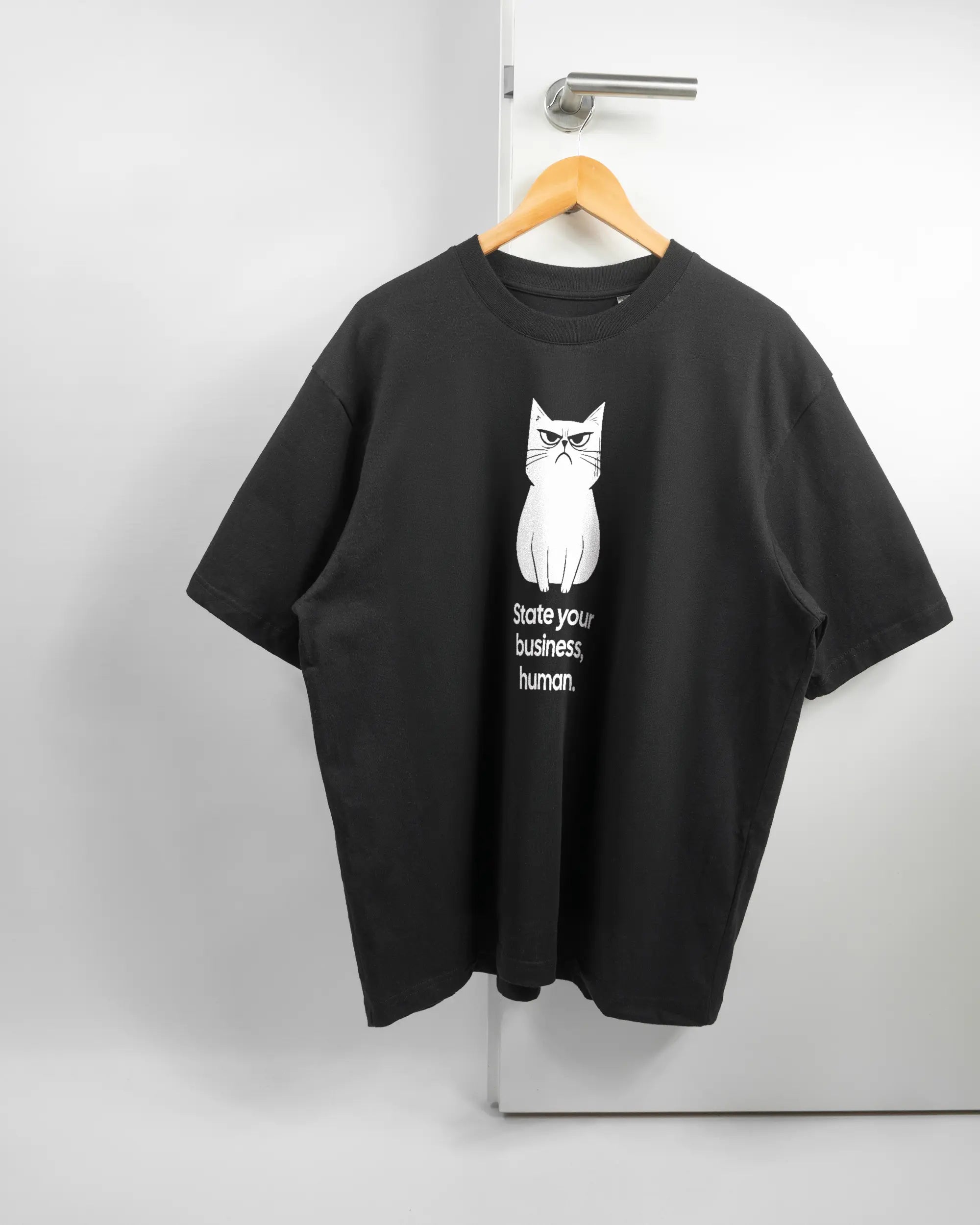 Vorderseite des Basic Unisex T-Shirt Oversized von Maondo mit dem Design Grumpy Purr in der Farbe Black und in der Größe L auf einem Bügel an der Türklinke aufgehängt