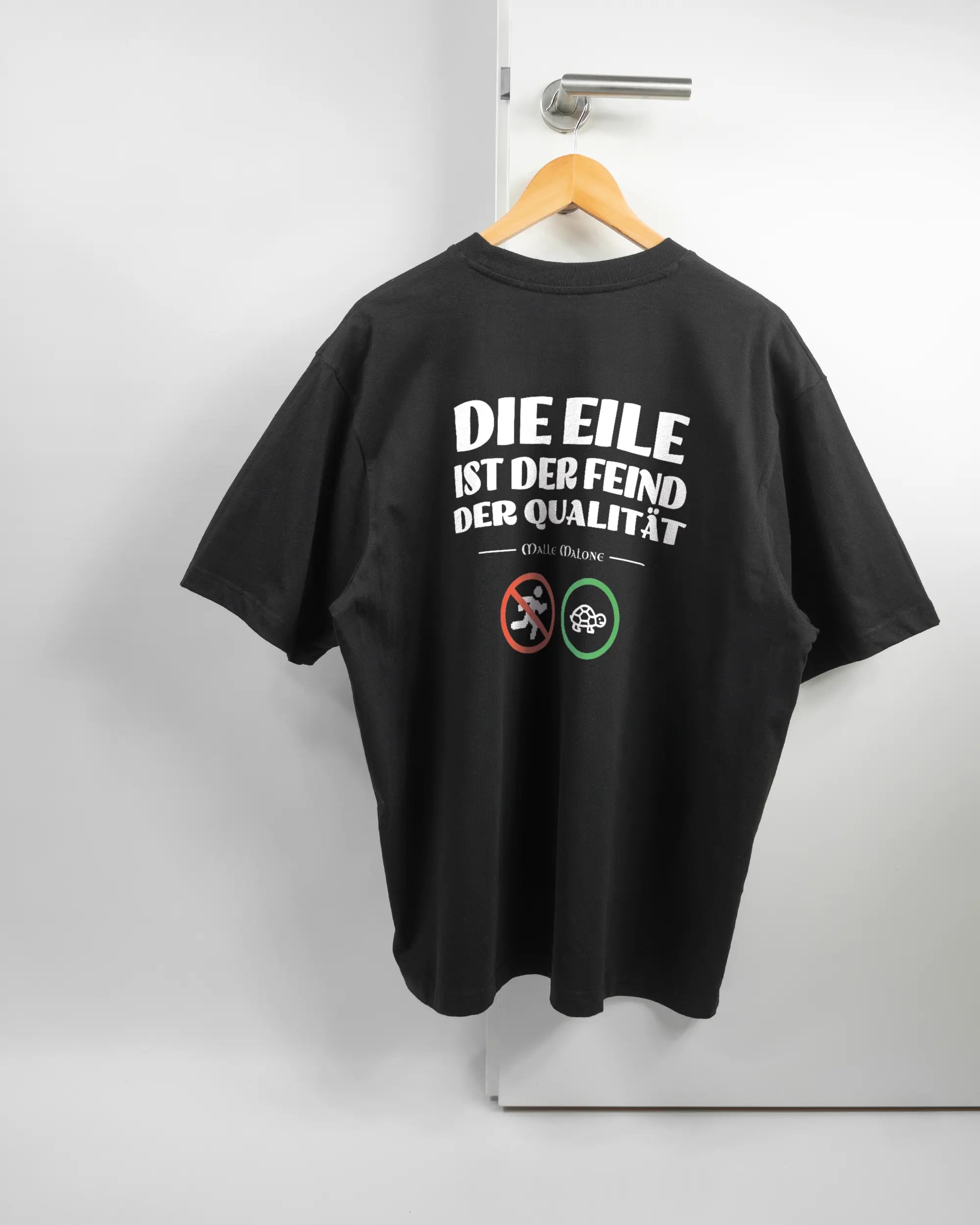 Rückseite des Basic Unisex T-Shirt Oversized von Maondo mit dem Design Keine Eile in der Farbe Black und in der Größe L auf einem Bügel an der Türklinke aufgehängt