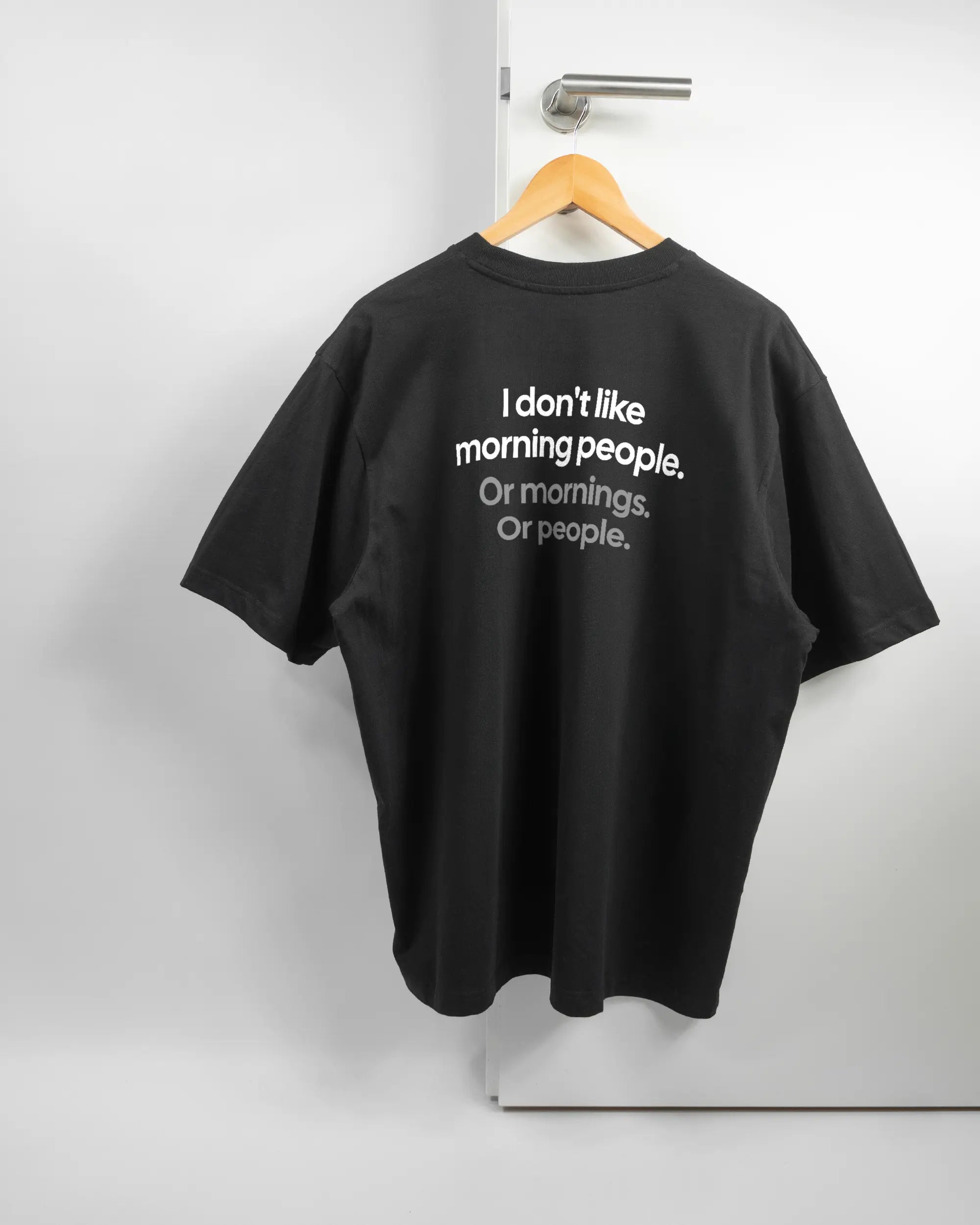 Rückseite des Basic Unisex T-Shirt Oversized von Maondo mit dem Design Morning Person in der Farbe Black und in der Größe L auf einem Bügel an der Türklinke aufgehängt
