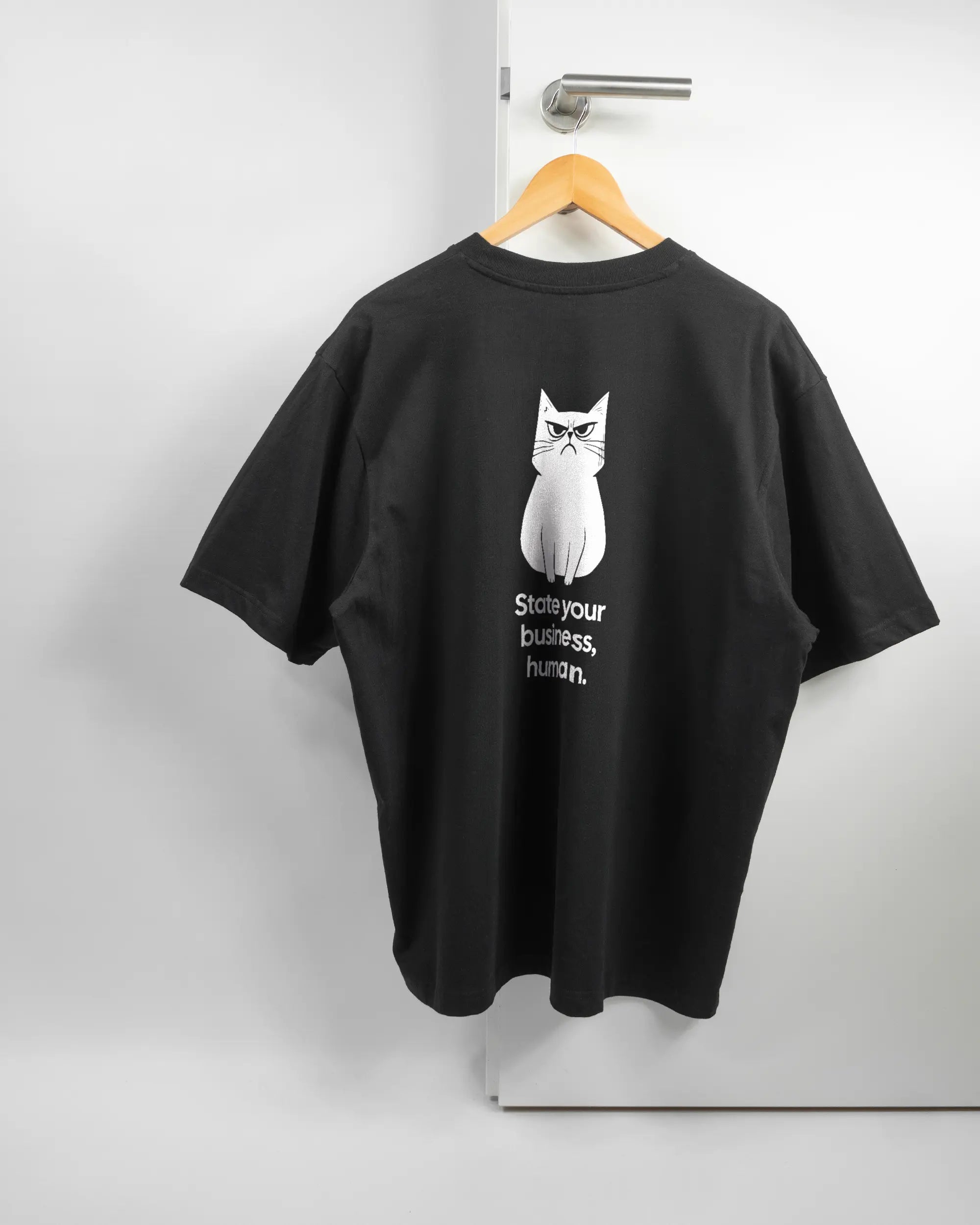 Rückseite des Basic Unisex T-Shirt Oversized von Maondo mit dem Design Grumpy Purr in der Farbe Black und in der Größe L auf einem Bügel an der Türklinke aufgehängt