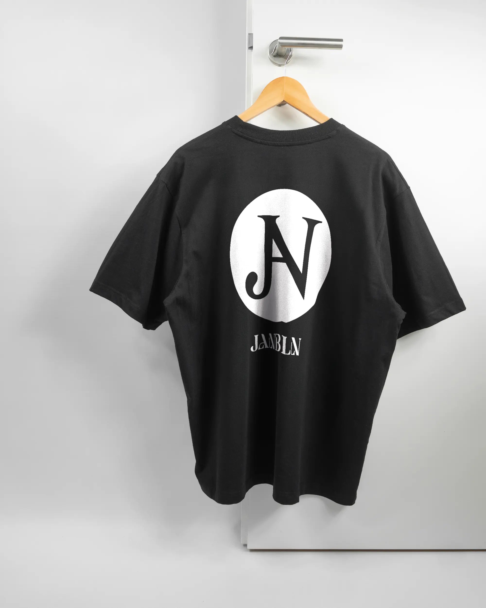 Rückseite des Basic Unisex T-Shirt Oversized von Maondo mit dem Design JB1 in der Farbe Black und in der Größe L auf einem Bügel an der Türklinke aufgehängt