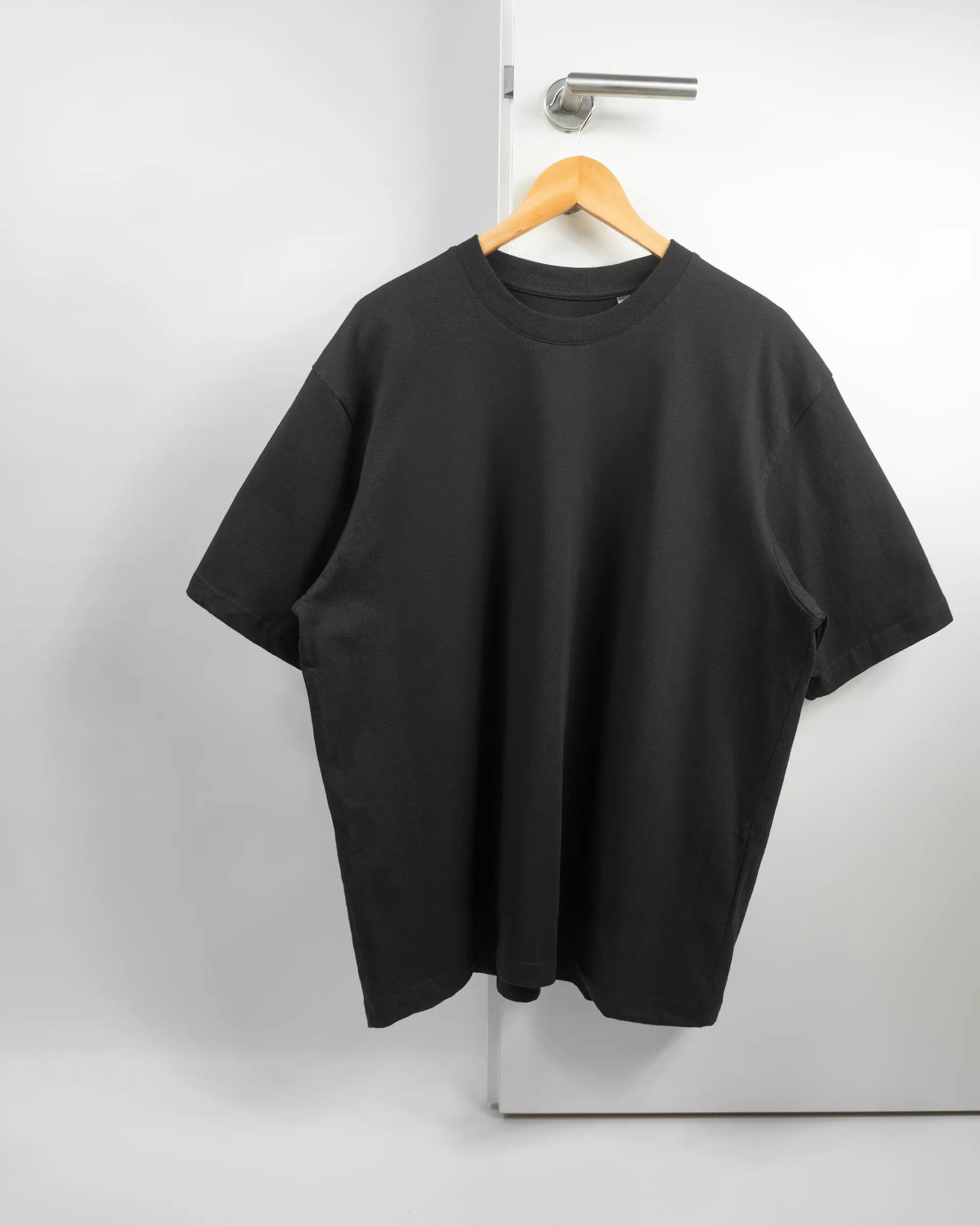 Vorderseite des Basic Unisex T-Shirt Oversized von Maondo ohne Motiv in der Farbe Black und in der Größe L auf einem Bügel an der Türklinke aufgehängt