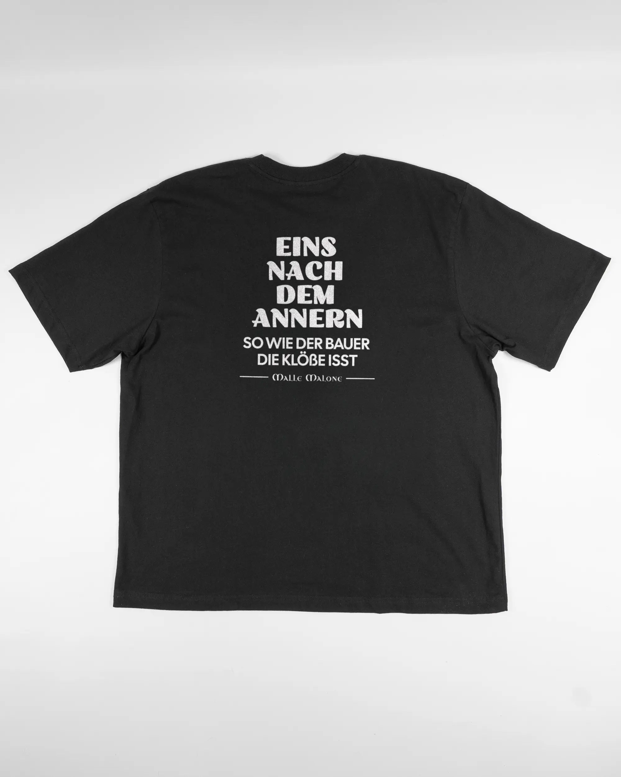 Rückseite des Basic Unisex T-Shirt Oversized von Maondo mit dem Design Eins nach dem Annern in der Farbe Black und in der Größe L flach auf den Boden gelegt